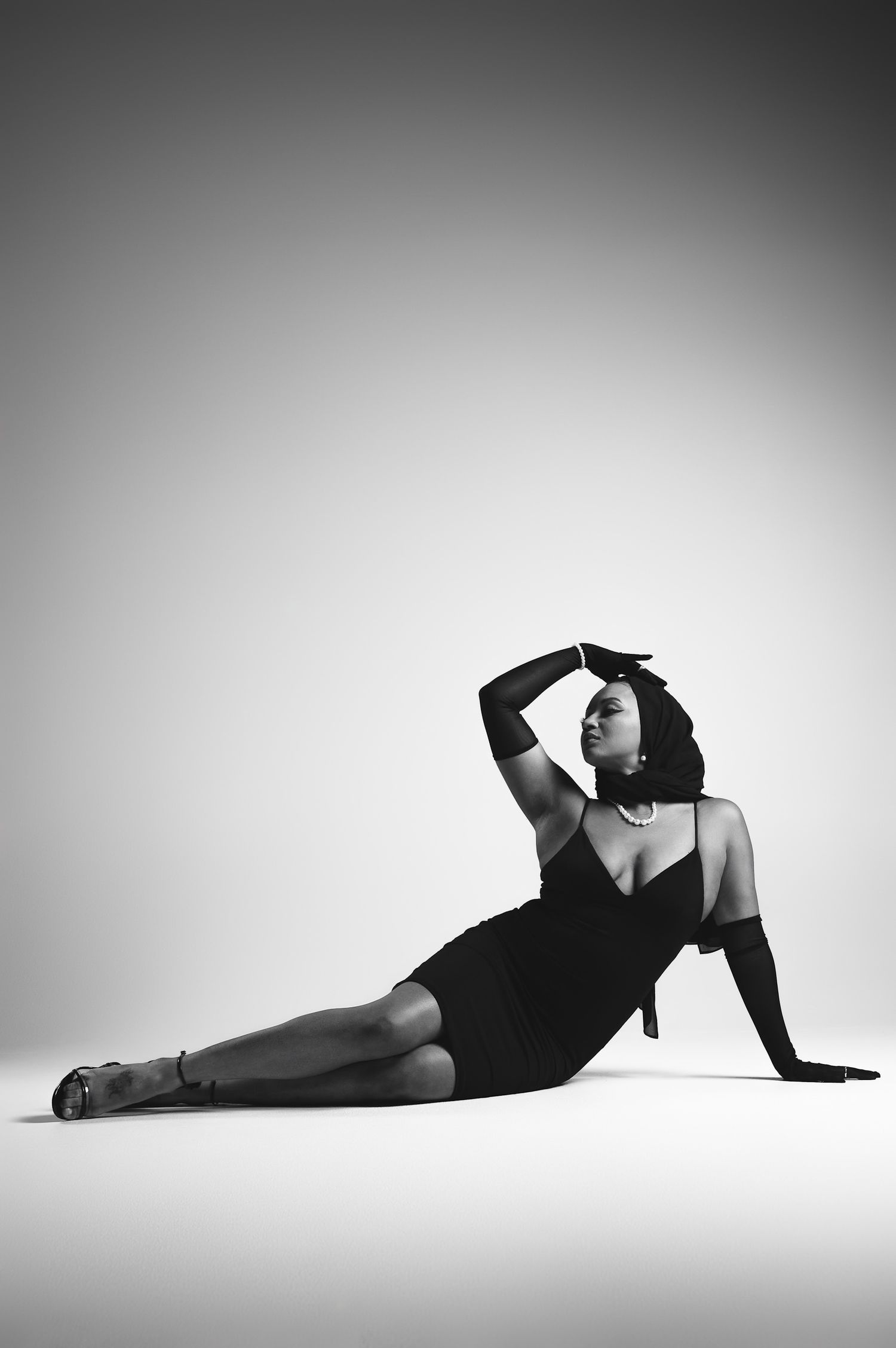 Black and white photo of a woman in a black dress and long gloves posing elegantly on the floor in a studio.