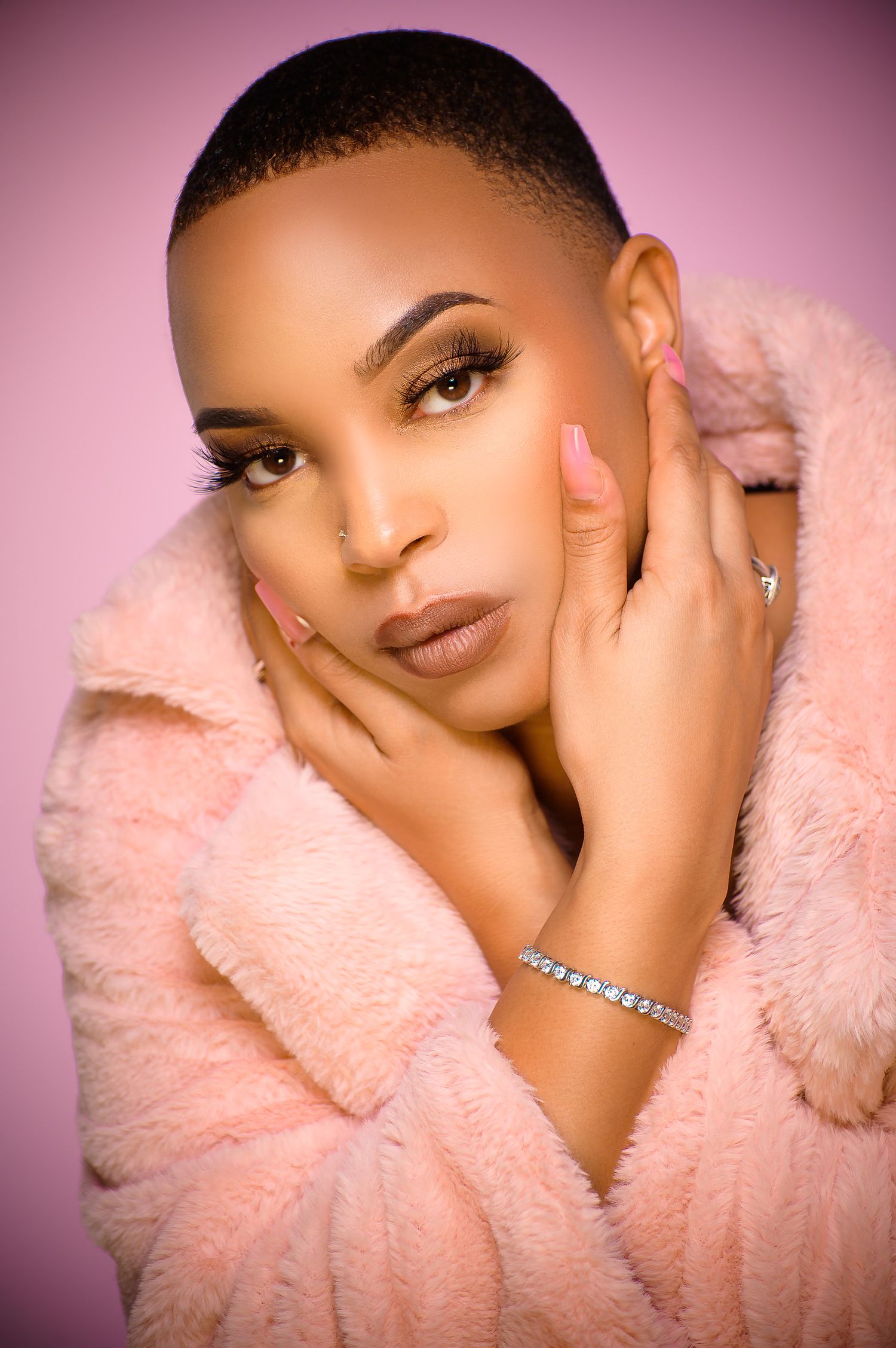 Glamorous woman in a pink fur coat with short hair, diamond bracelet, and pink nails on pink background.