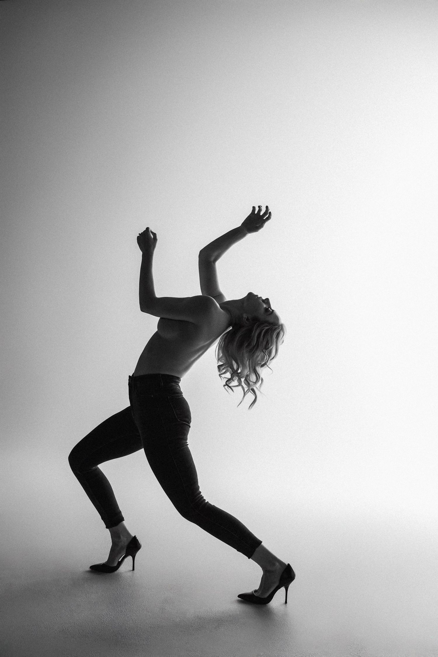 Black and white photo of a woman dancing dynamically in heels and jeans, arms raised expressively against a light background.