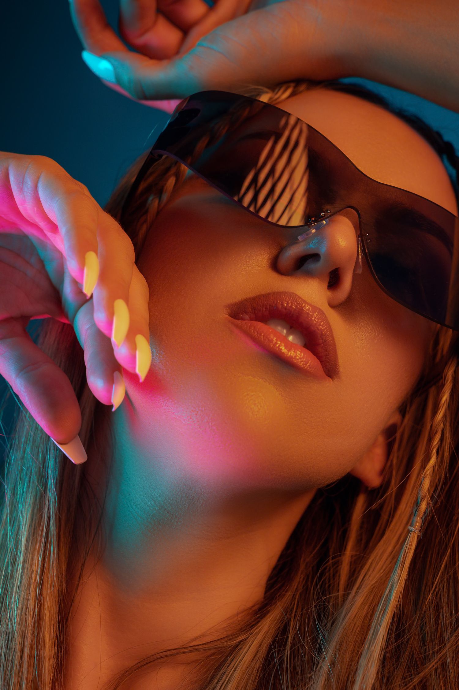 Woman in dark sunglasses with neon pink and teal lighting, glossy lips, and colorful nails in a fashion portrait.