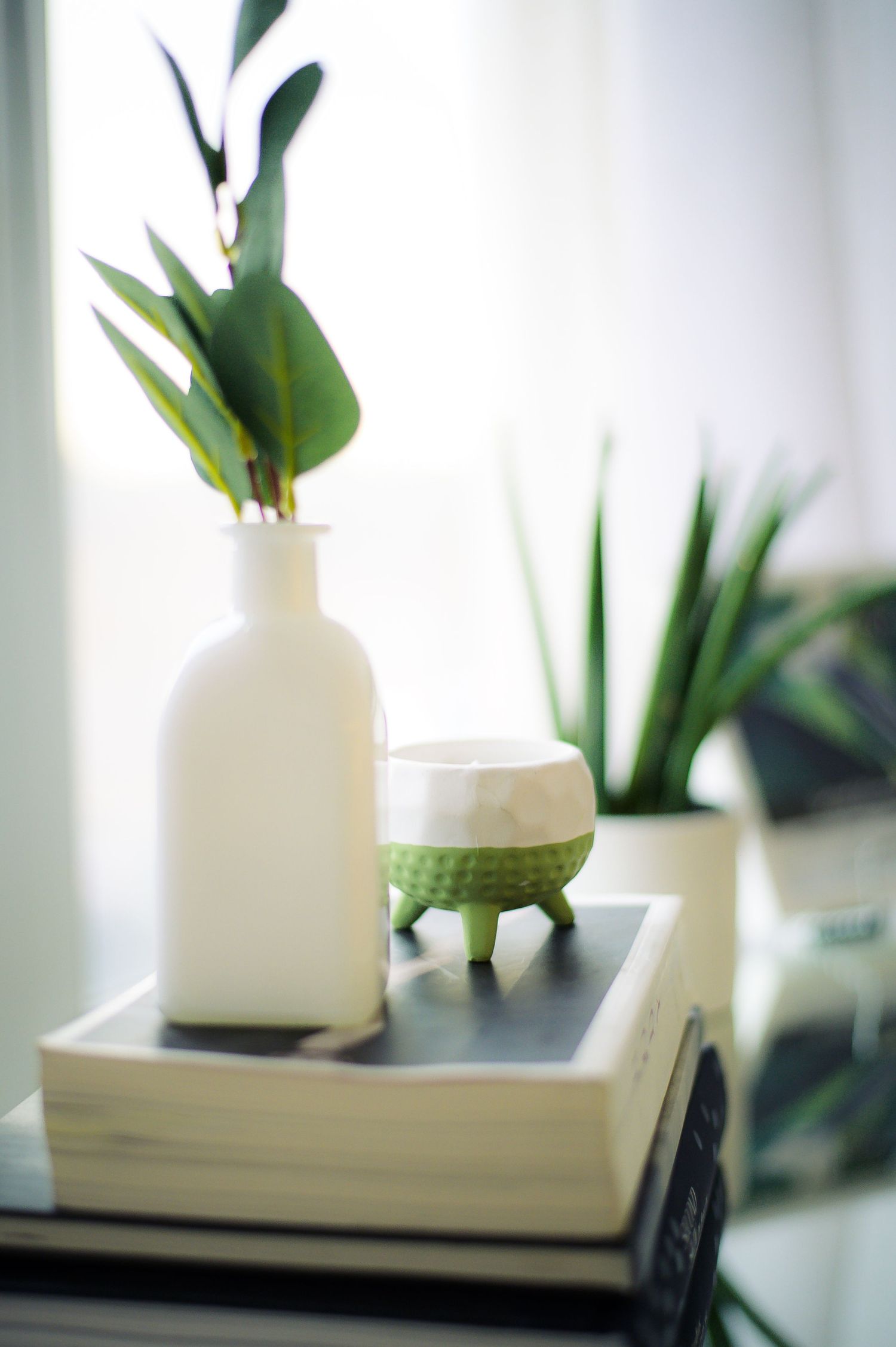 White ceramic vase with green plant leaves sits on a modern white surface near a window with soft natural lighting.