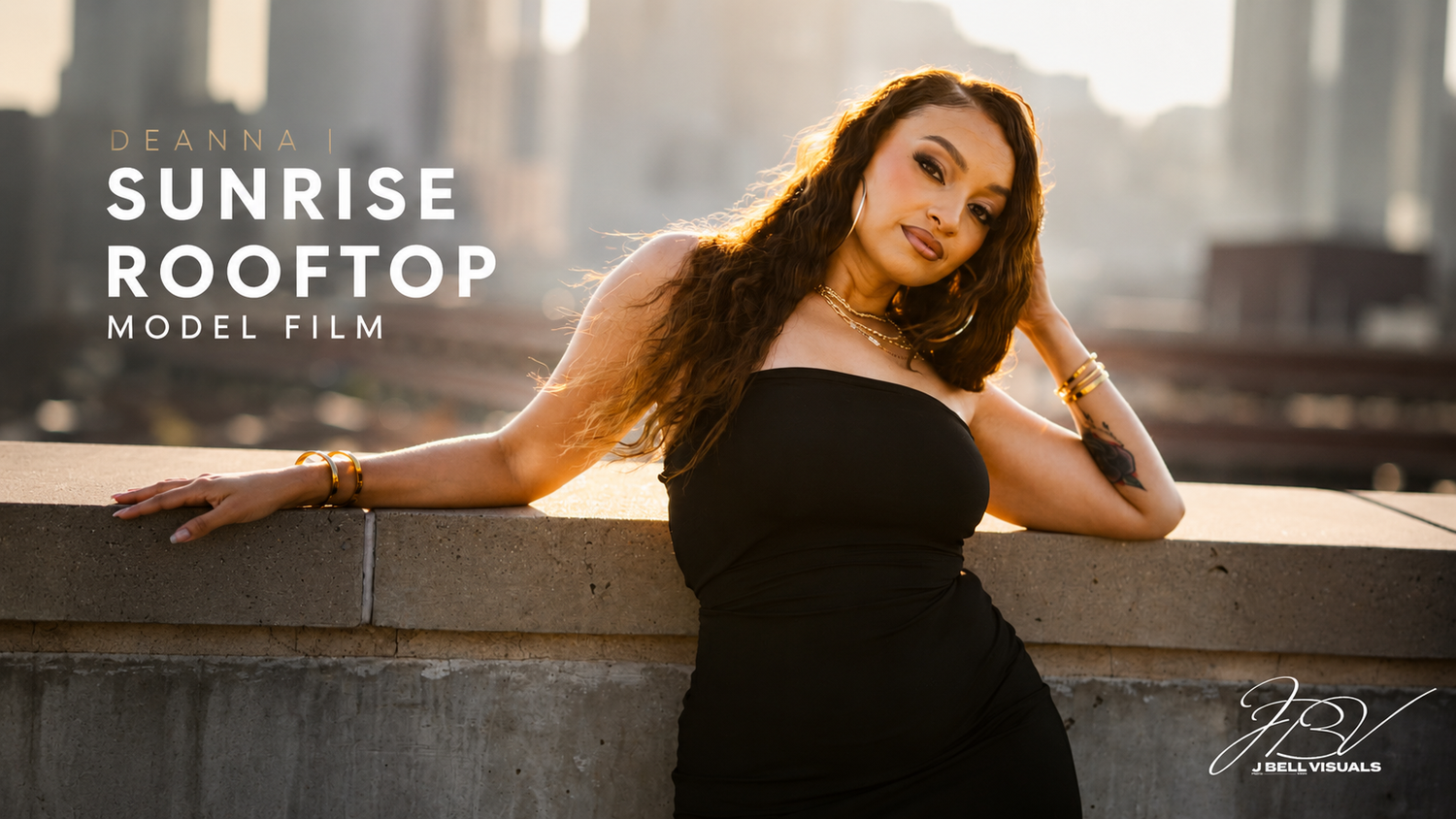 A woman in a black strapless dress poses on a rooftop at sunrise with a city skyline in the background.