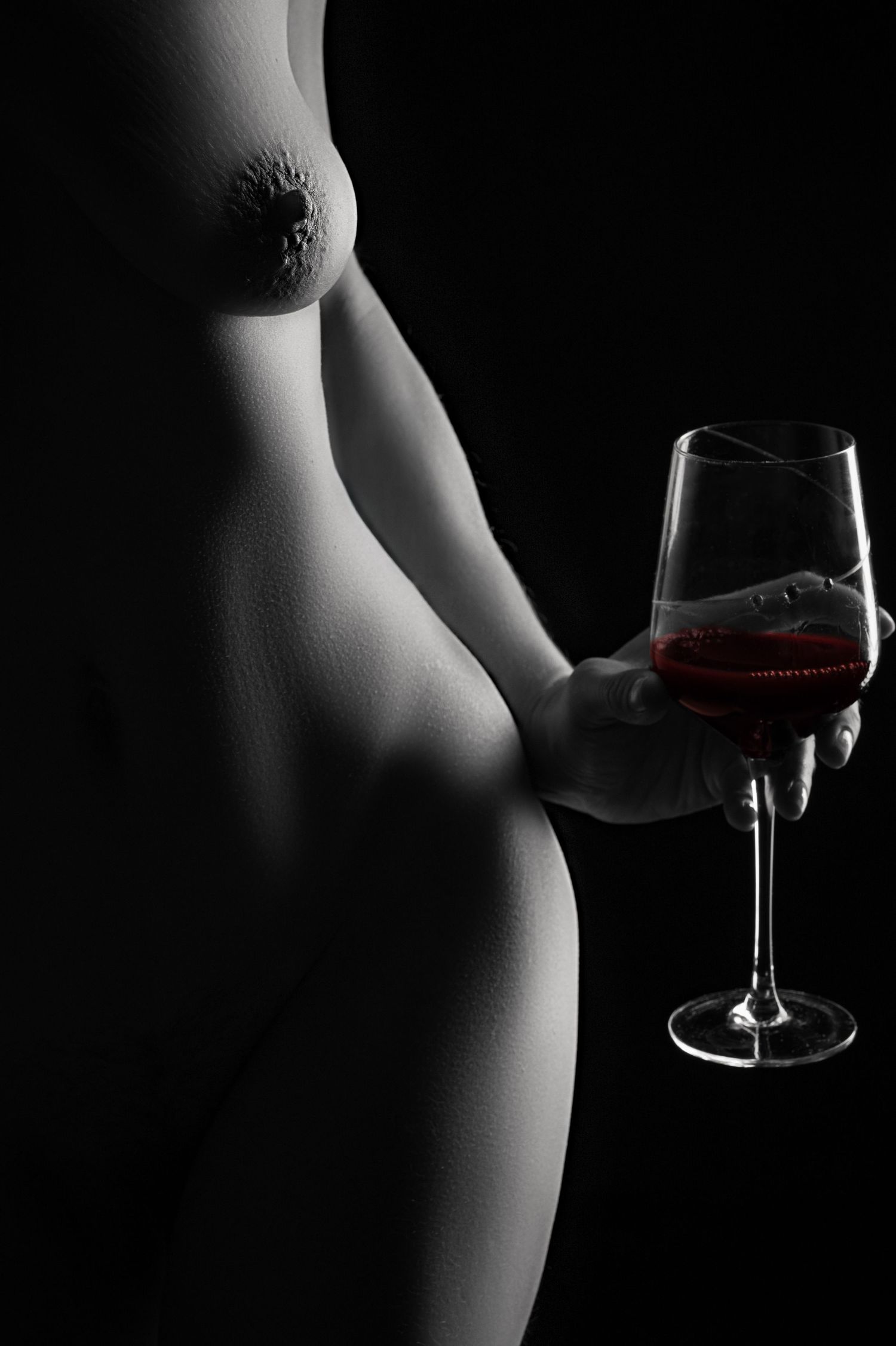 Black and white artistic nude figure holding a red wine glass against dark background.