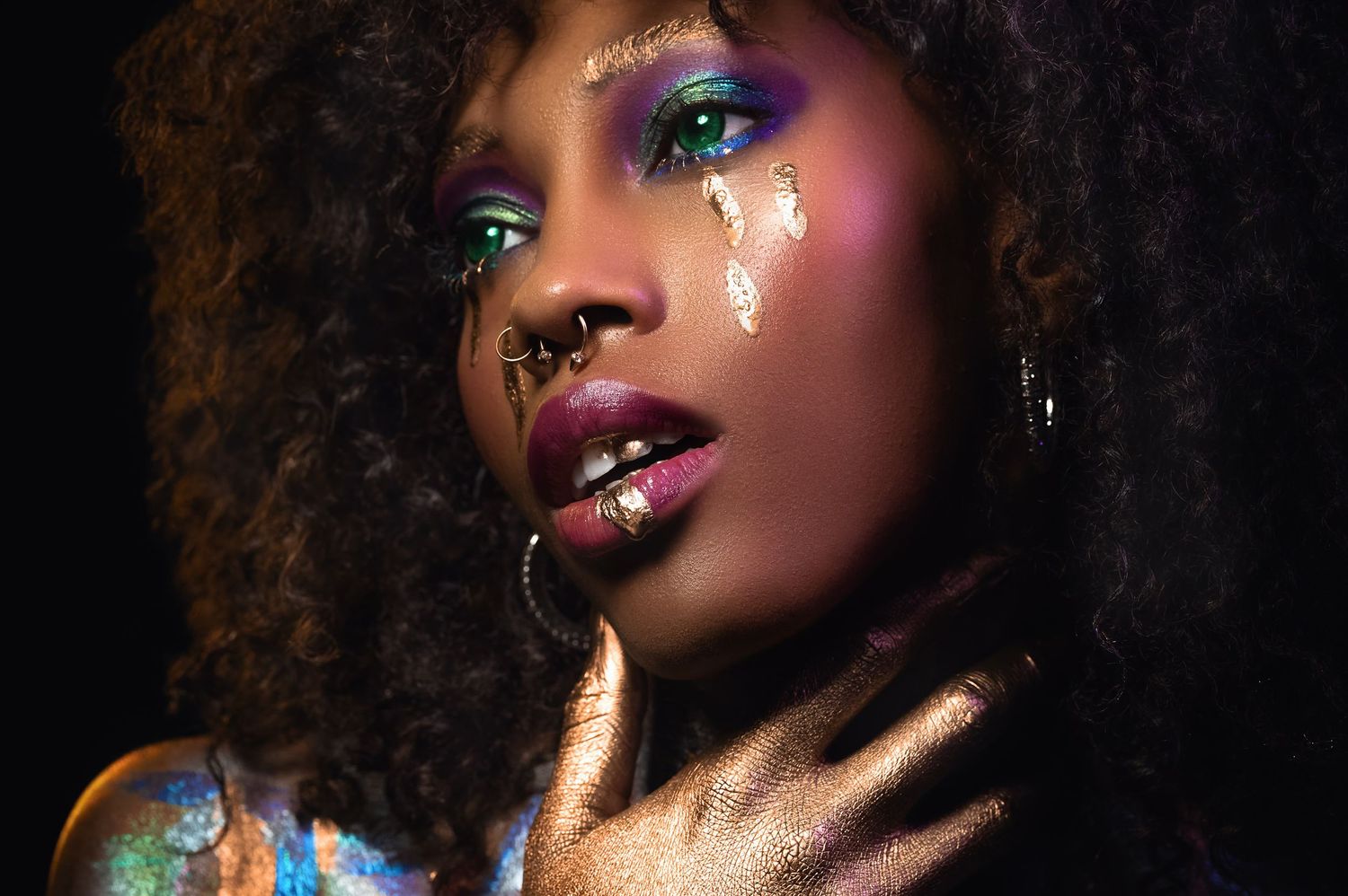 Close-up portrait of a woman with vibrant colorful makeup, glitter tears, and afro hair against dark background.