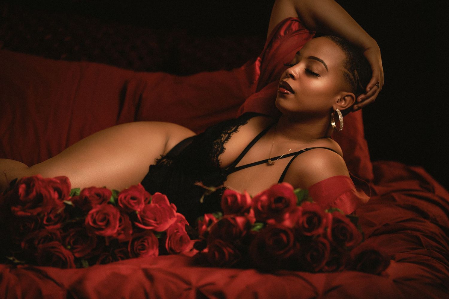 A romantic boudoir photo session with red roses and dramatic lighting against dark burgundy bedding.