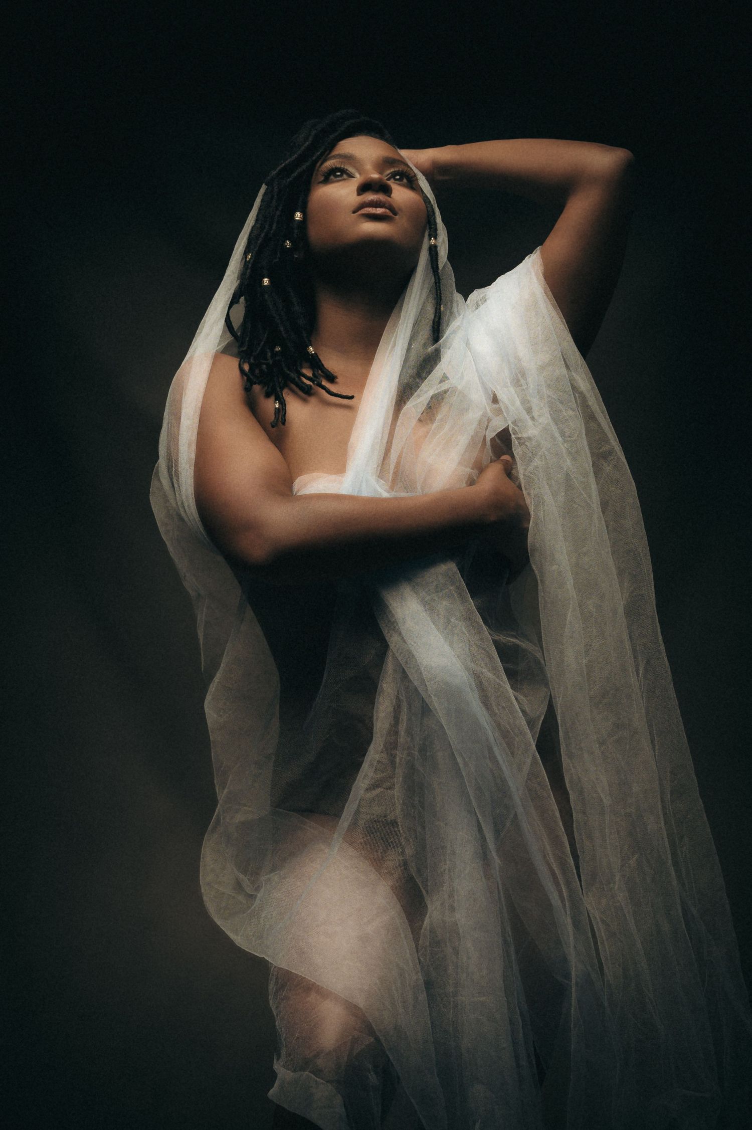 A Black woman draped in flowing white sheer fabric poses gracefully against a dark studio background.