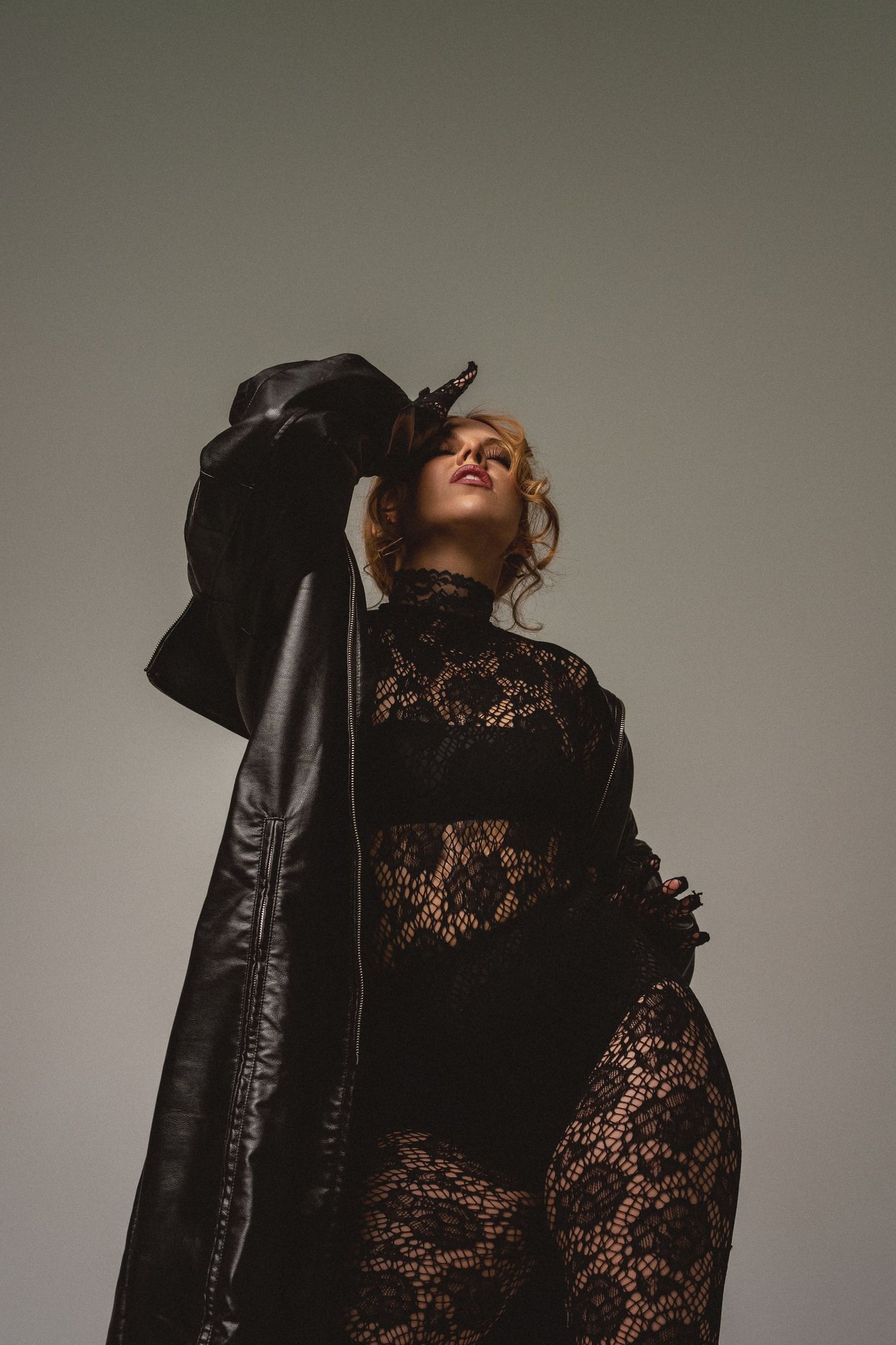 Woman in black lace bodysuit and leather jacket posing dramatically against gray studio background.
