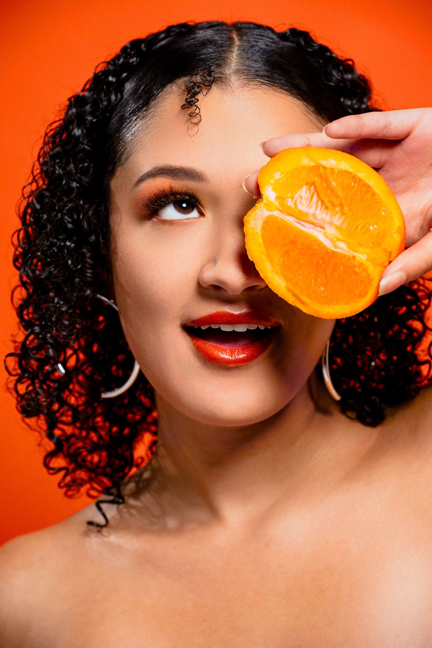 Smiling woman with curly hair holding a halved orange over her eye against a vibrant orange background.