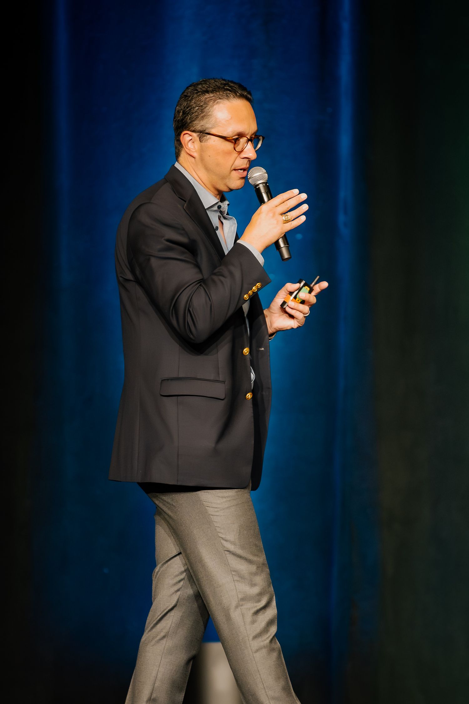 Man in blazer speaking into microphone on stage, holding a clicker during a professional presentation.