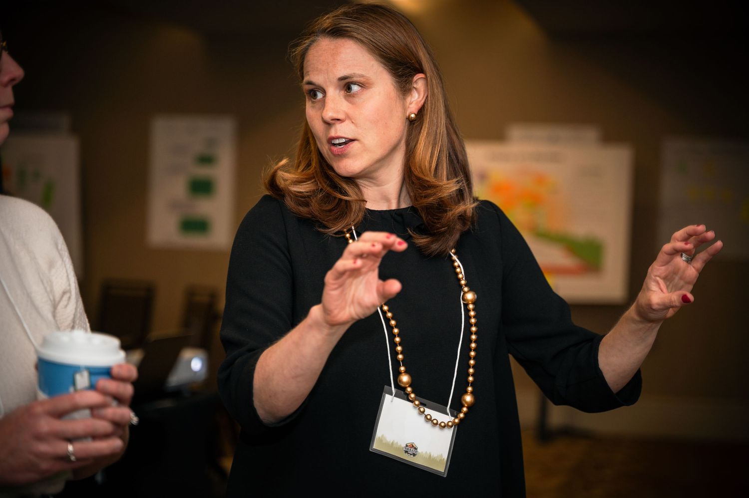 Woman in black top with pearl necklace and lanyard gestures expressively during a professional conference event.