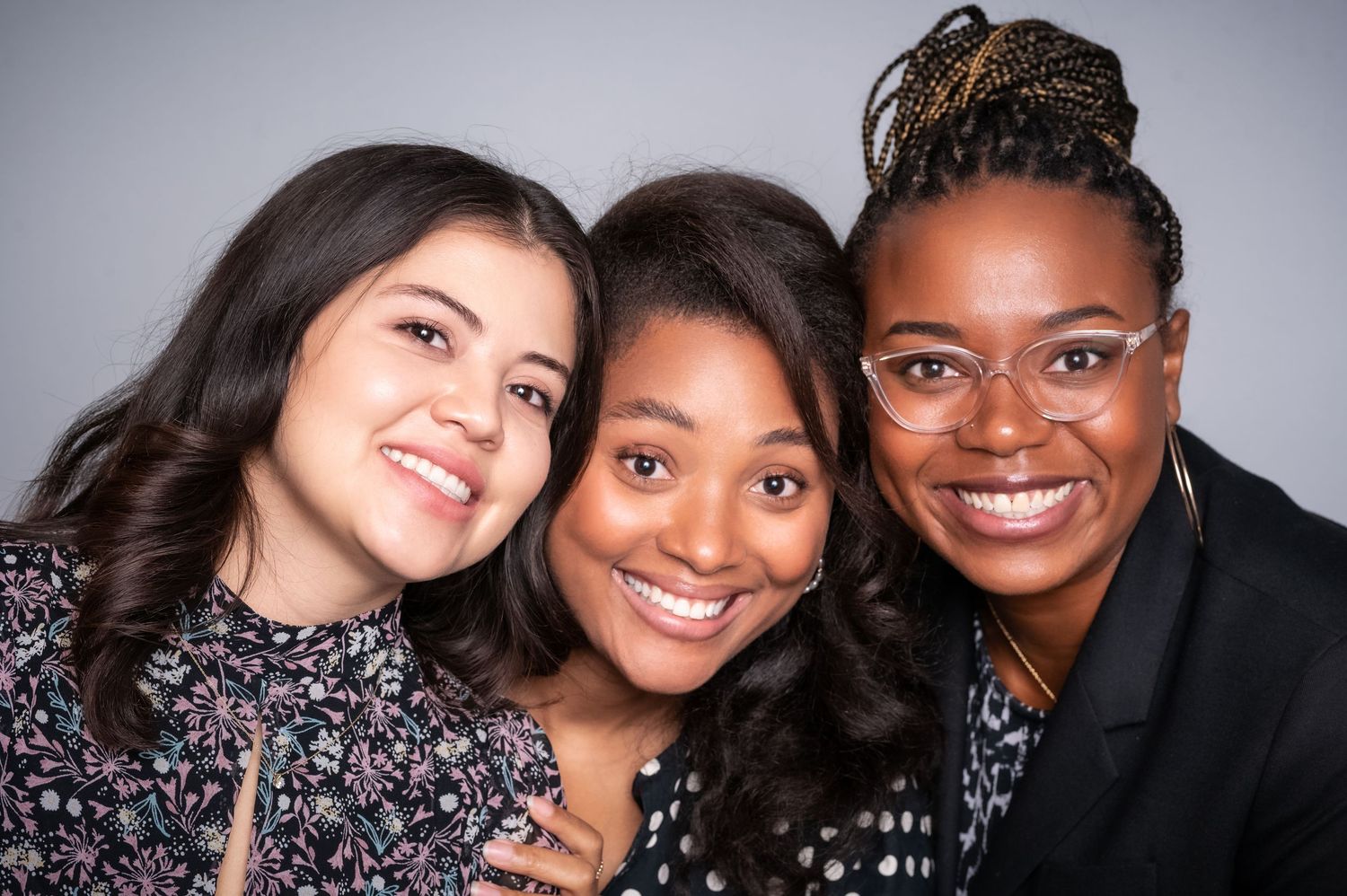 Three diverse friends smile warmly together in a casual studio portrait against a gray background.
