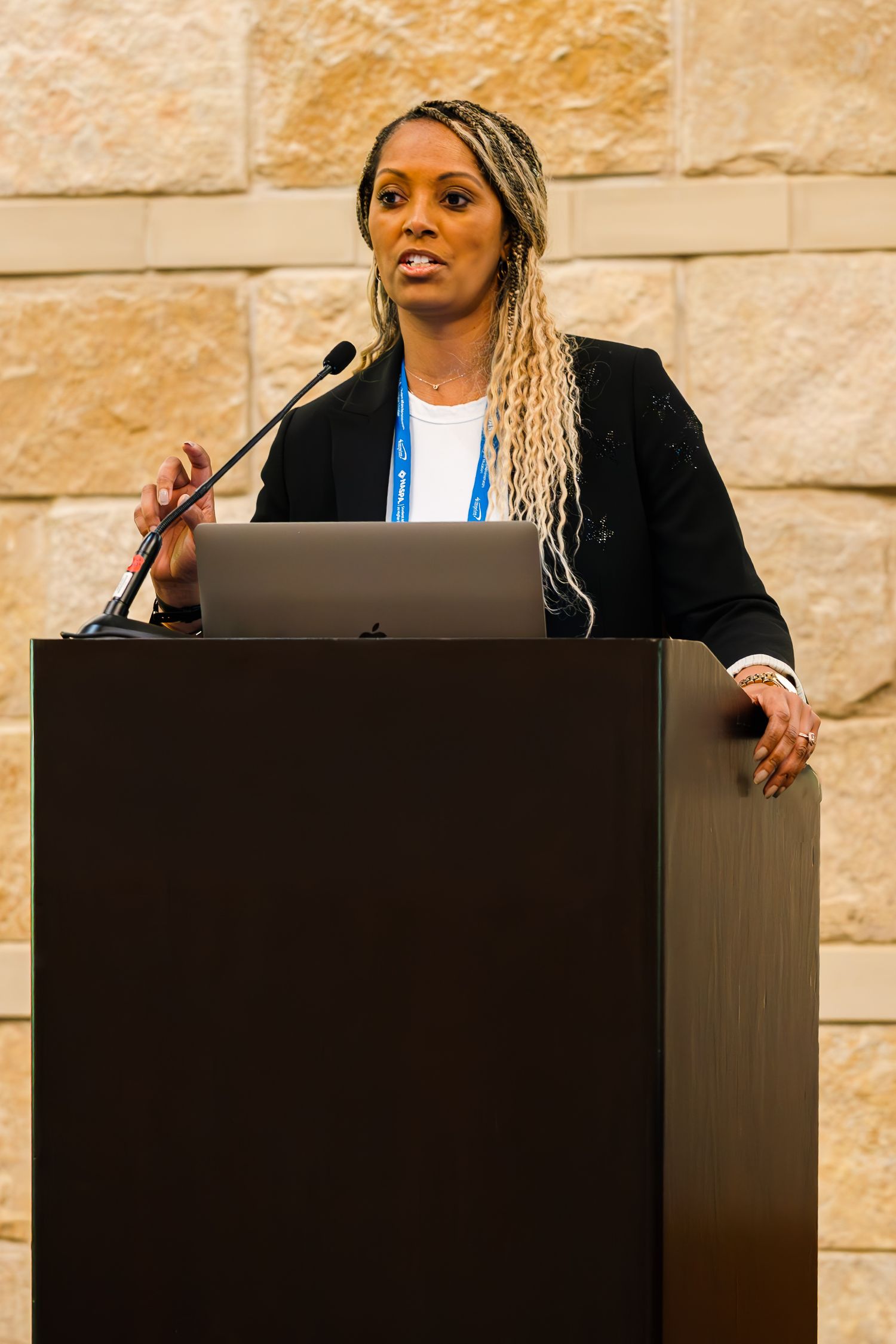 Woman with braided hair speaks at a podium during a professional conference, wearing a black blazer and blue lanyard.