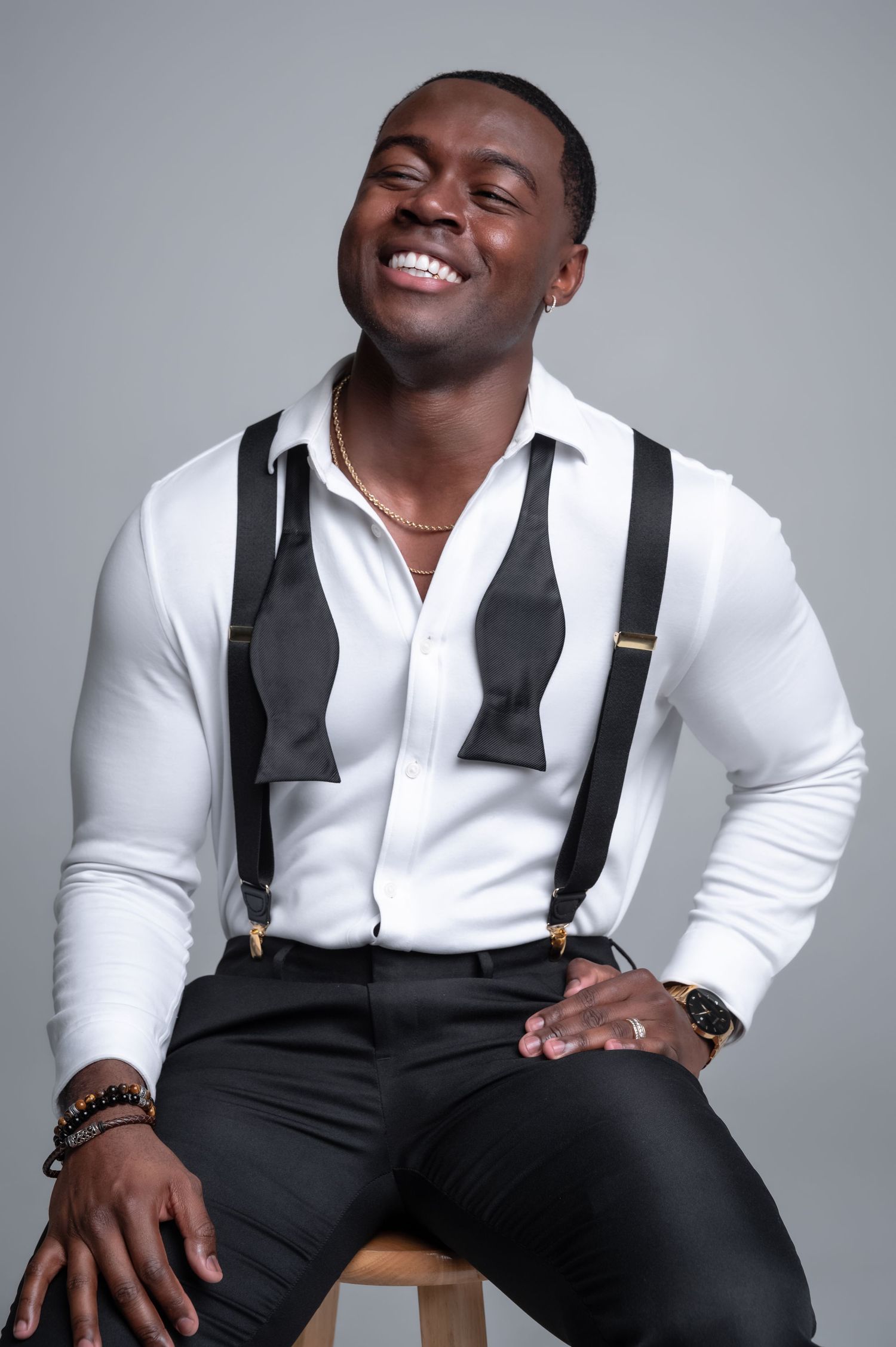 A smiling person in a crisp white dress shirt and black suspenders sits casually on a stool against a gray backdrop.