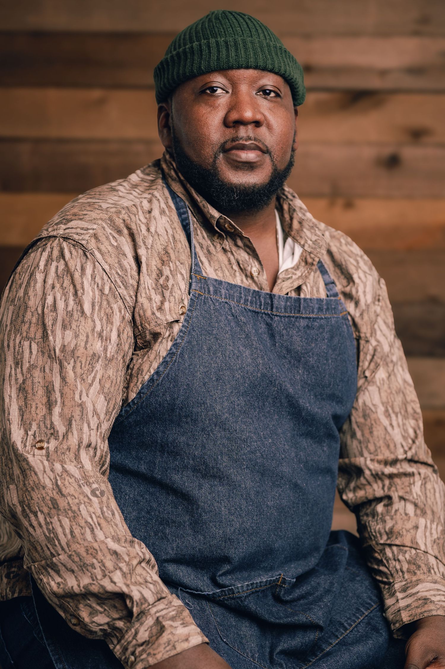 Casual portrait in denim apron, knit beanie and textured sweater against wooden wall.