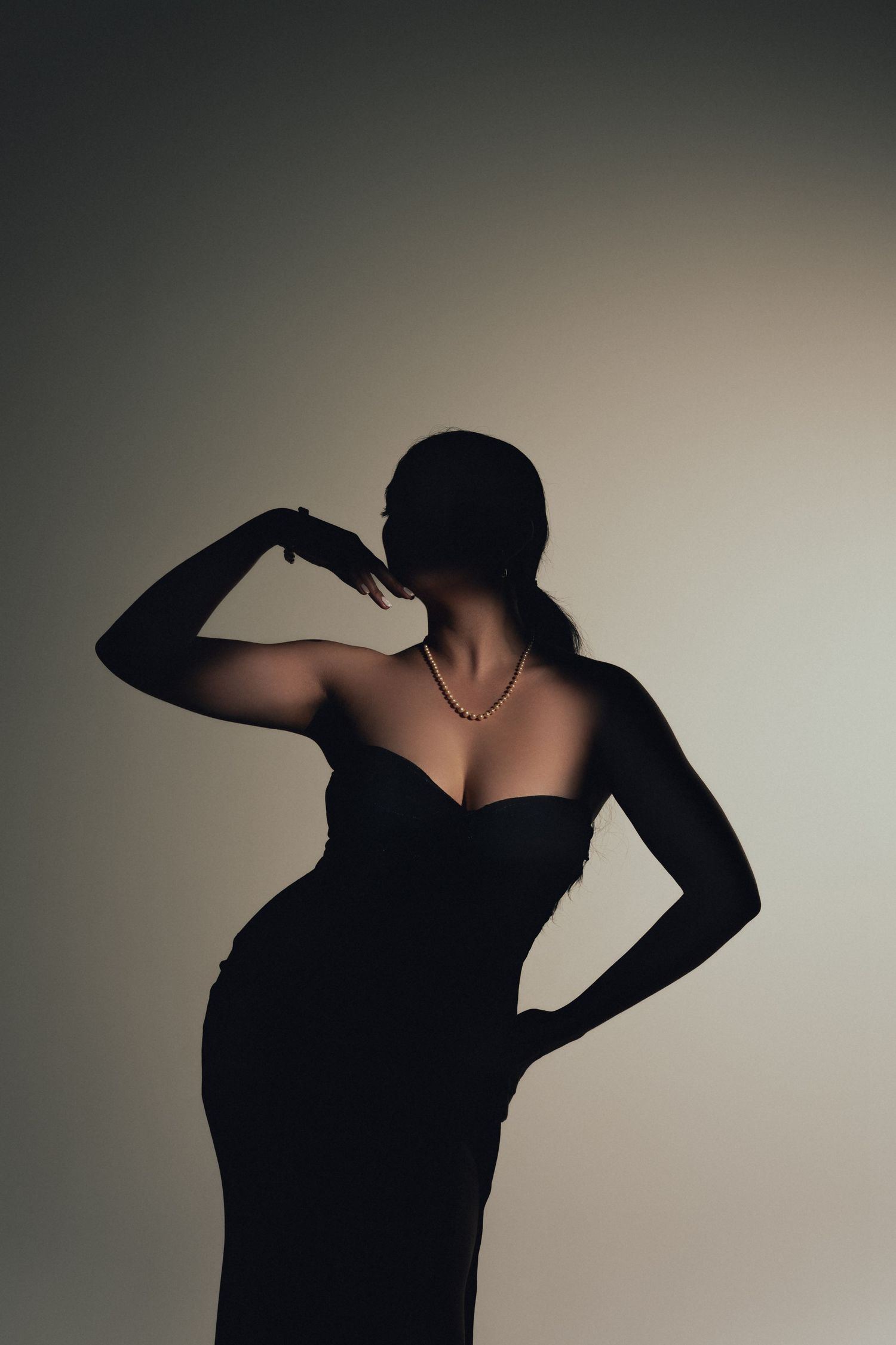 Elegant silhouette in black evening gown poses against neutral background with dramatic side lighting.