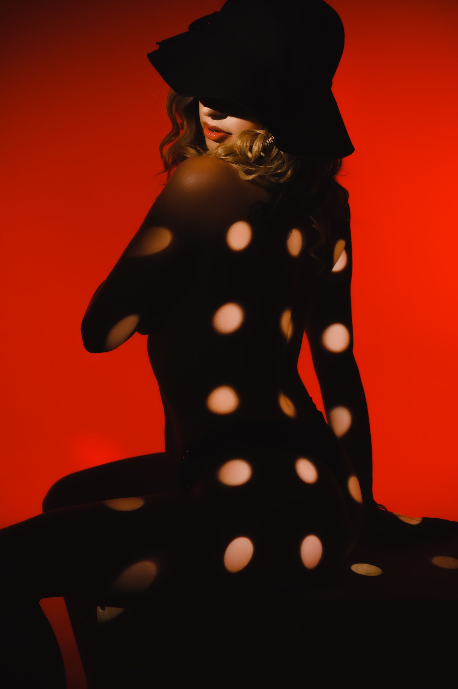 A woman in a black hat sits against a red background with circular light spots projected across her body.