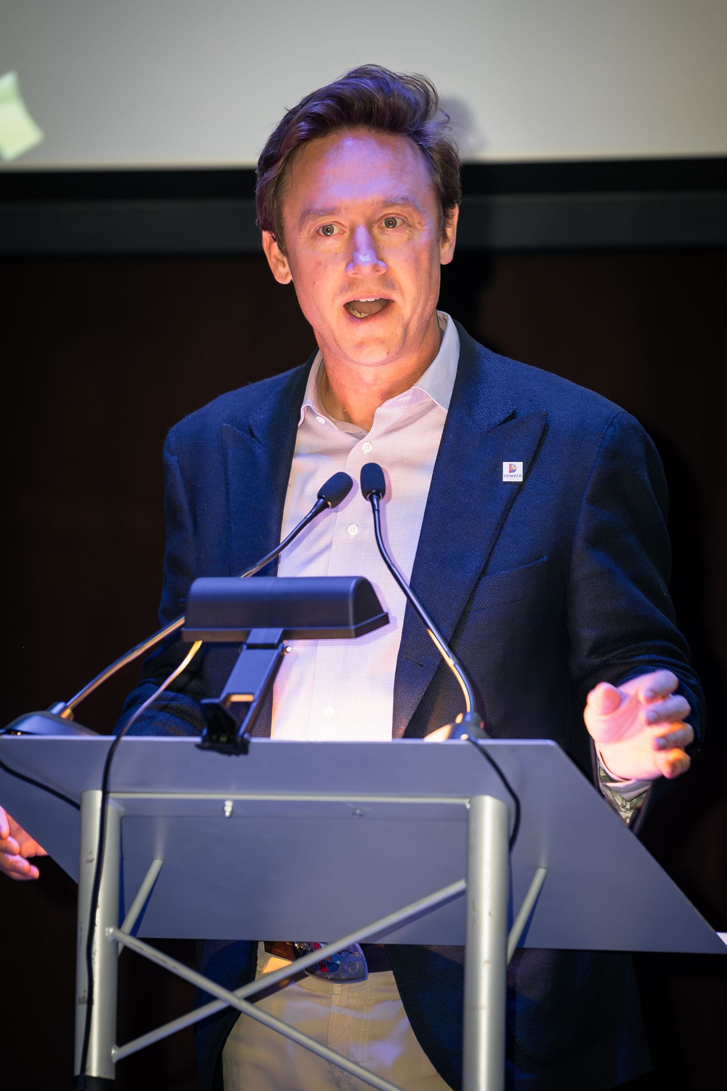 Denver, CO Mayor Mike Johnston in blue blazer speaking at a podium with microphones during a professional conference event.