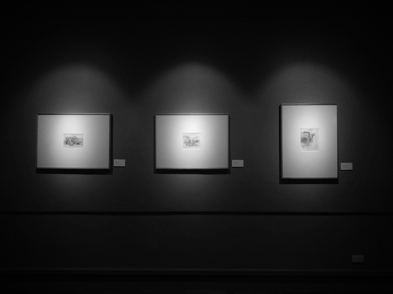 Monochromatic art gallery wall displays minimalist framed photographs under spotlights in a dark room.