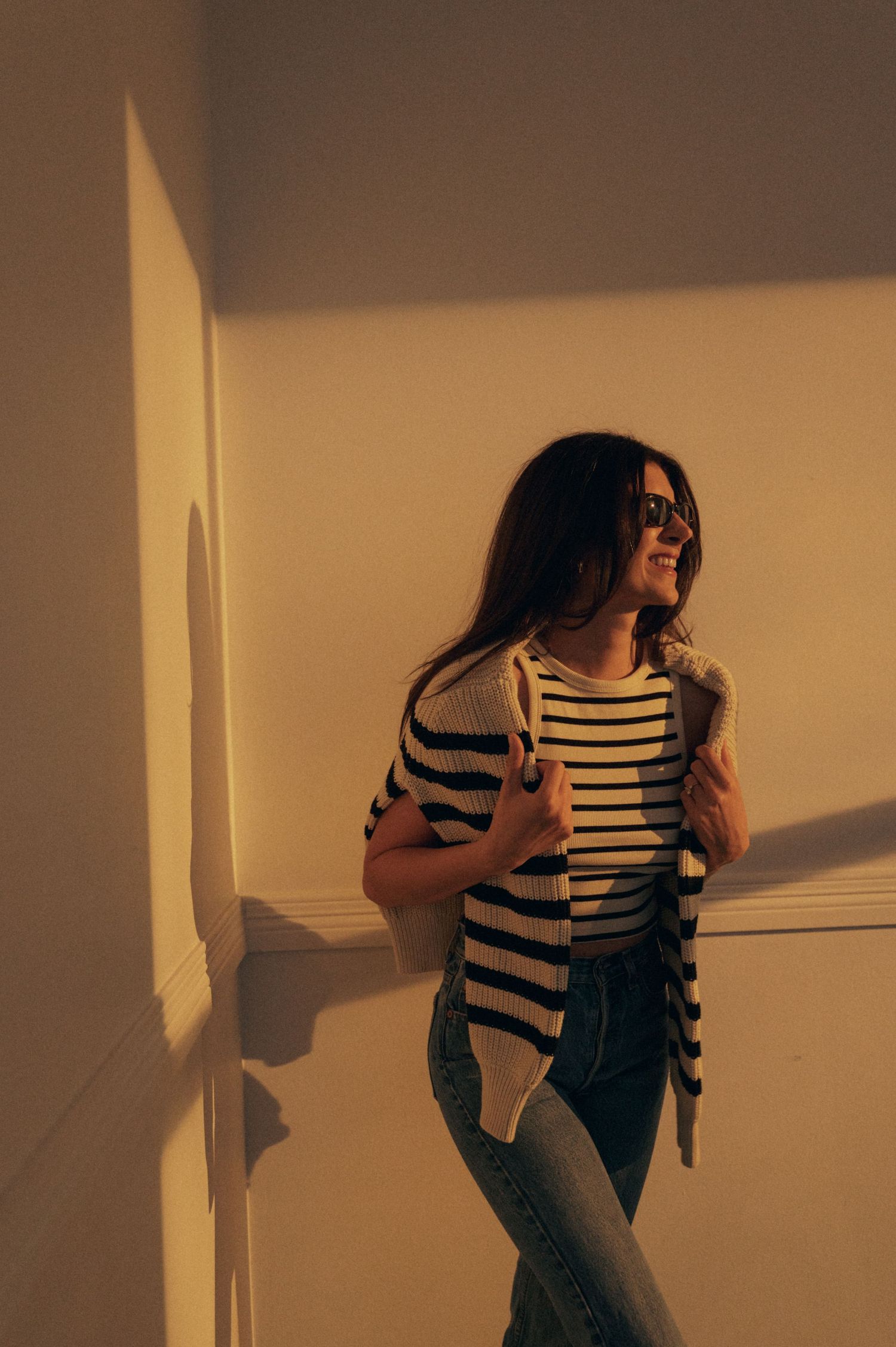 Person in striped shirt and jeans leaning against wall in warm sunset lighting.