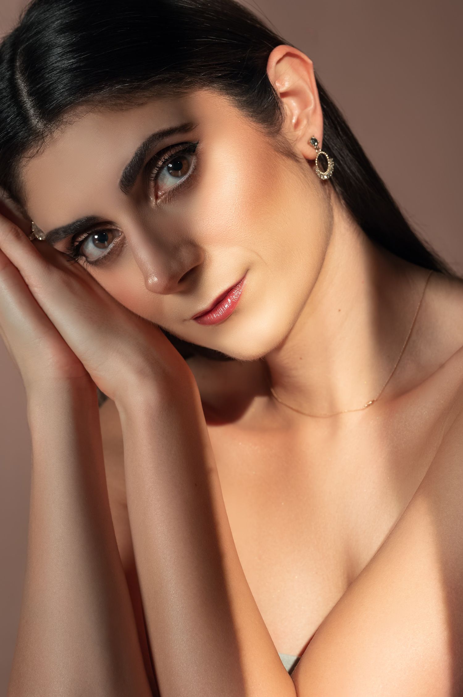 Young woman with dark hair wearing gold earrings and delicate necklace, posing gracefully with hands near face.