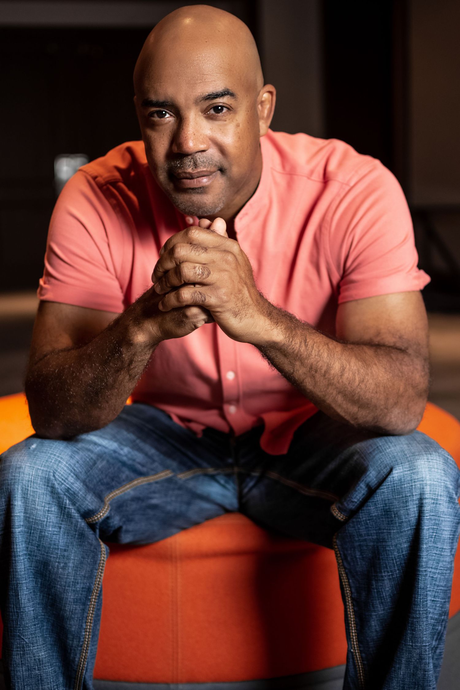 Man in coral pink shirt poses on orange chair during professional photo shoot.
