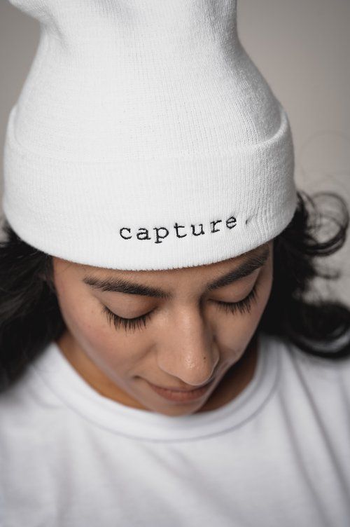 A person wearing a white beanie with the word 'capture' embroidered on it looks down with closed eyes.