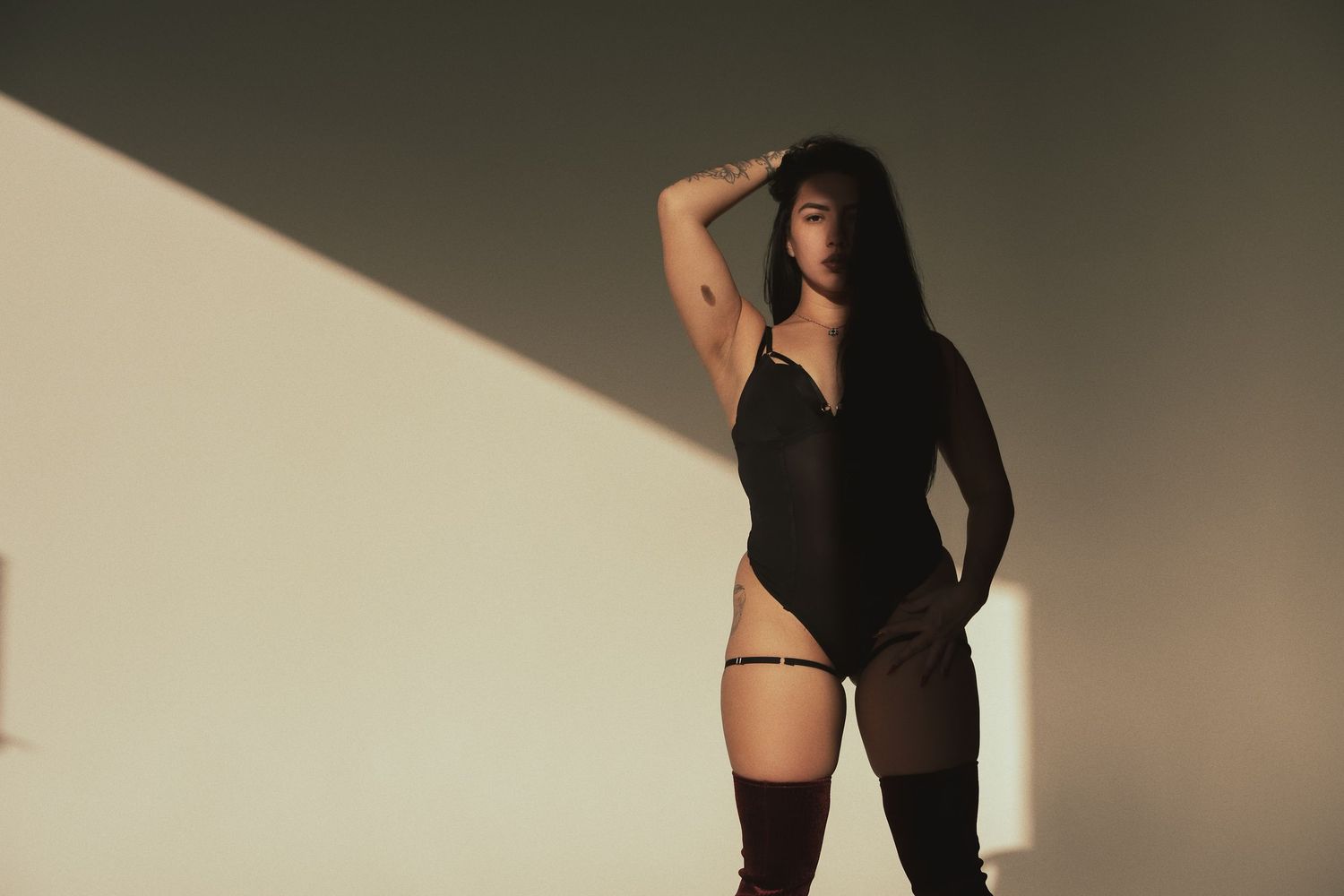 Woman in black bodysuit and thigh-high stockings posing in dramatic natural light against a neutral wall.