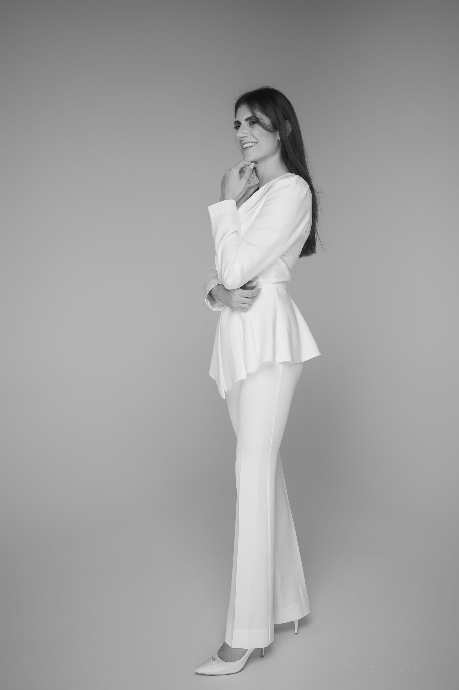 Woman in white peplum blazer and wide-leg trousers poses elegantly in black and white fashion portrait.