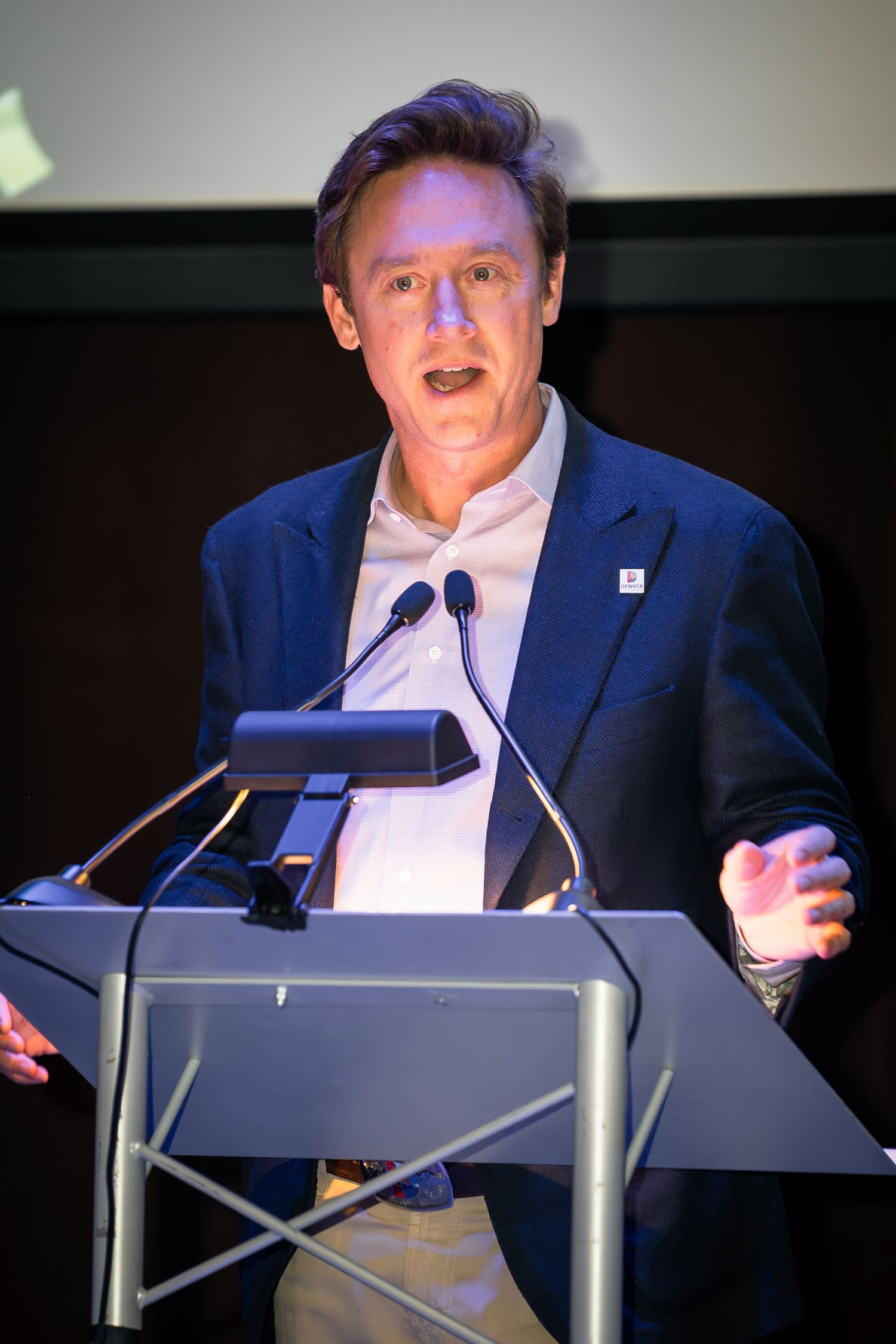 Man in blue blazer speaking at a podium with microphones during a professional conference event.