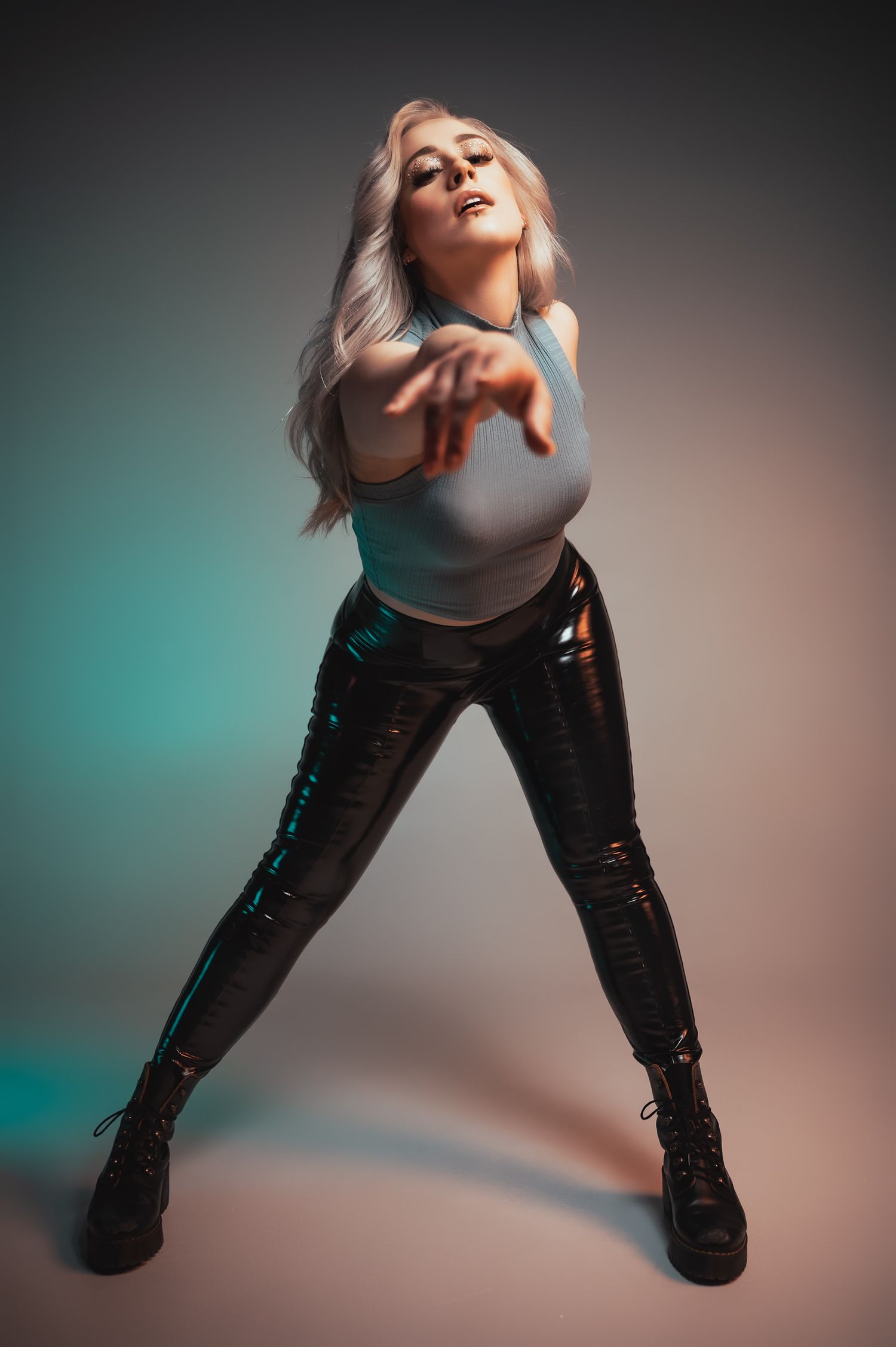 Woman in shiny black leggings and white top poses dynamically under colorful studio lighting with blonde hair.