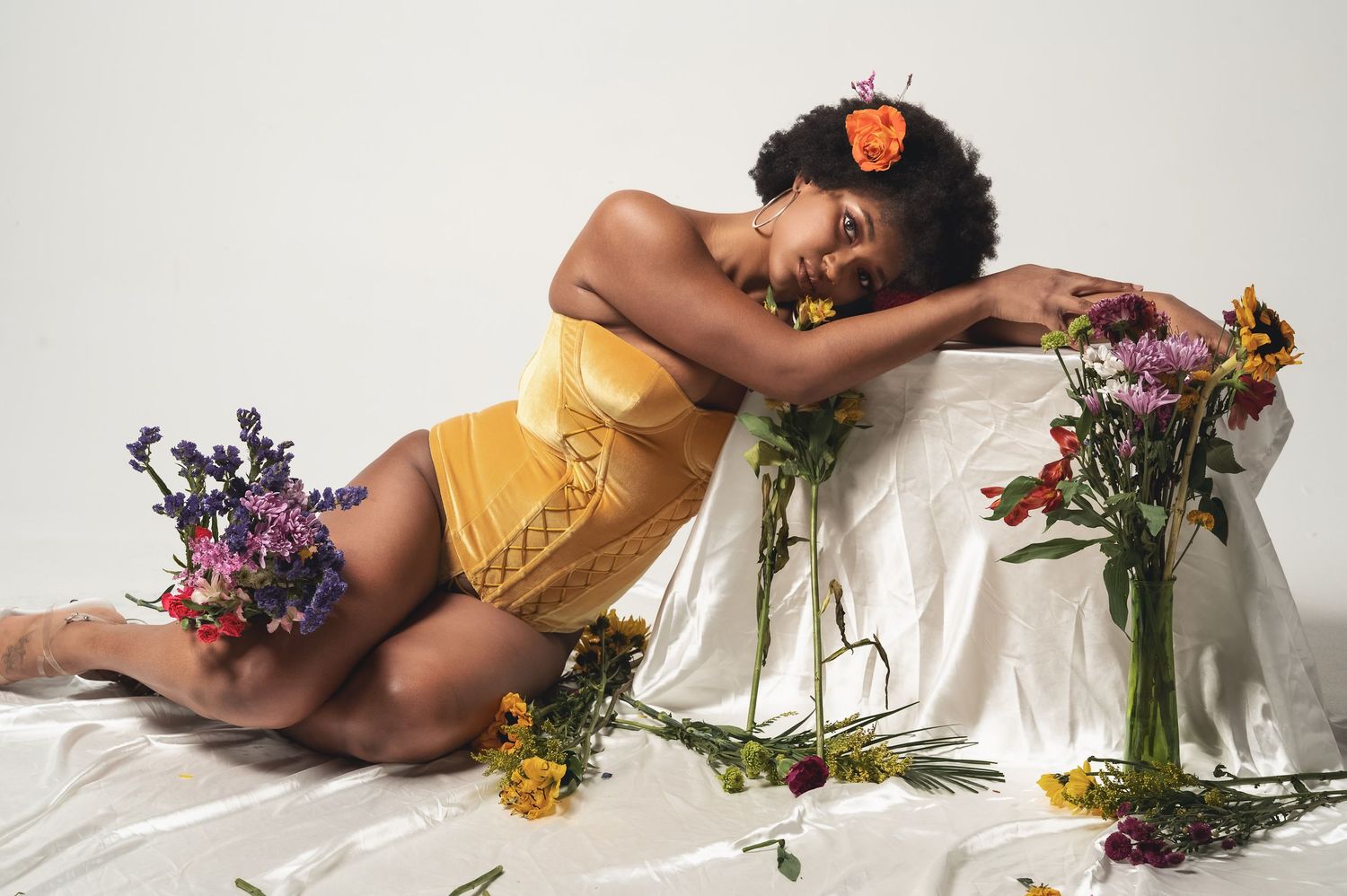 Woman in yellow outfit poses with colorful wildflowers on white fabric, orange flower in natural hair.
