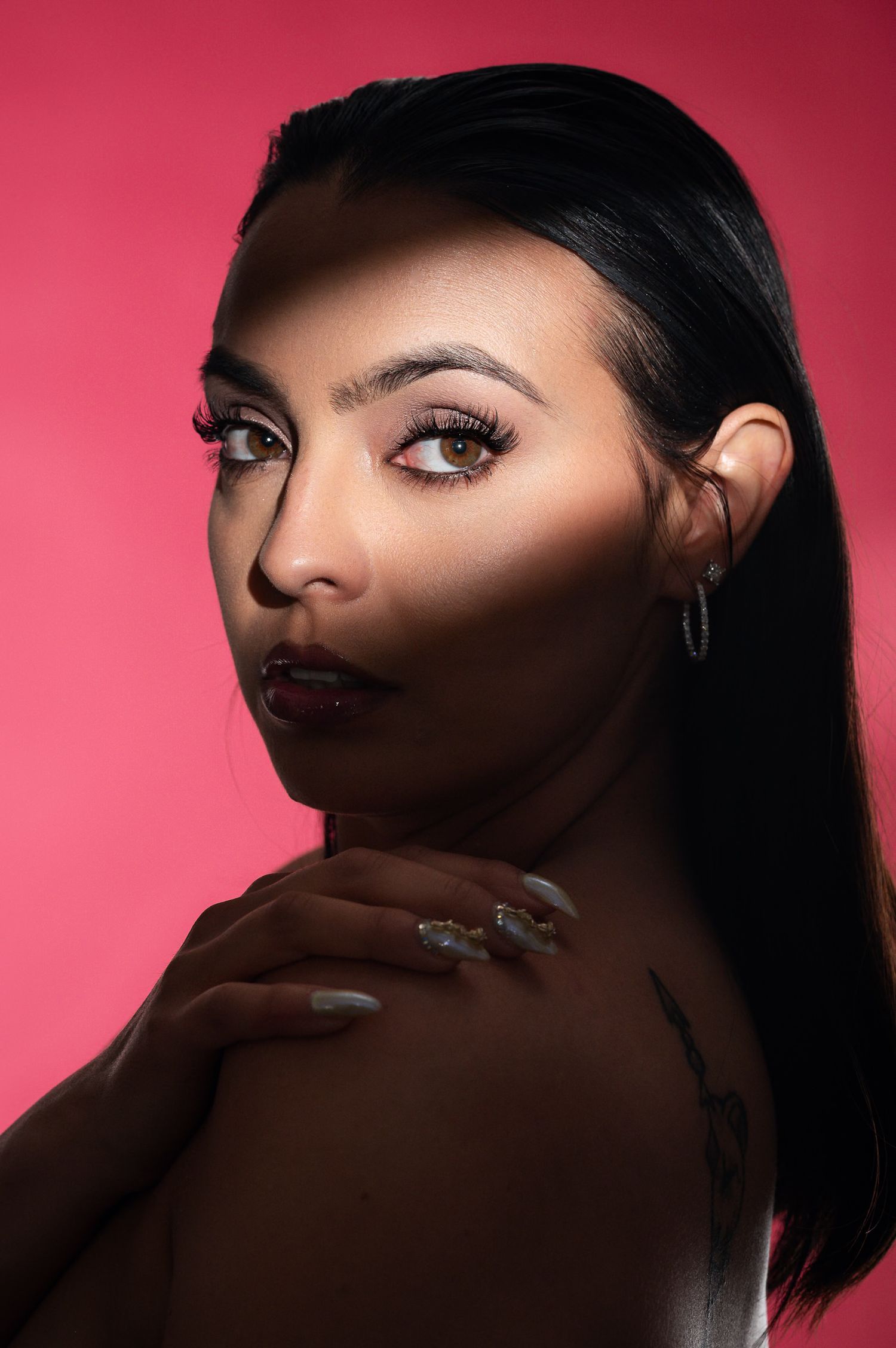 A woman with bold makeup, dark lips, and styled black hair poses against a vibrant pink background, showcasing elegant nails.