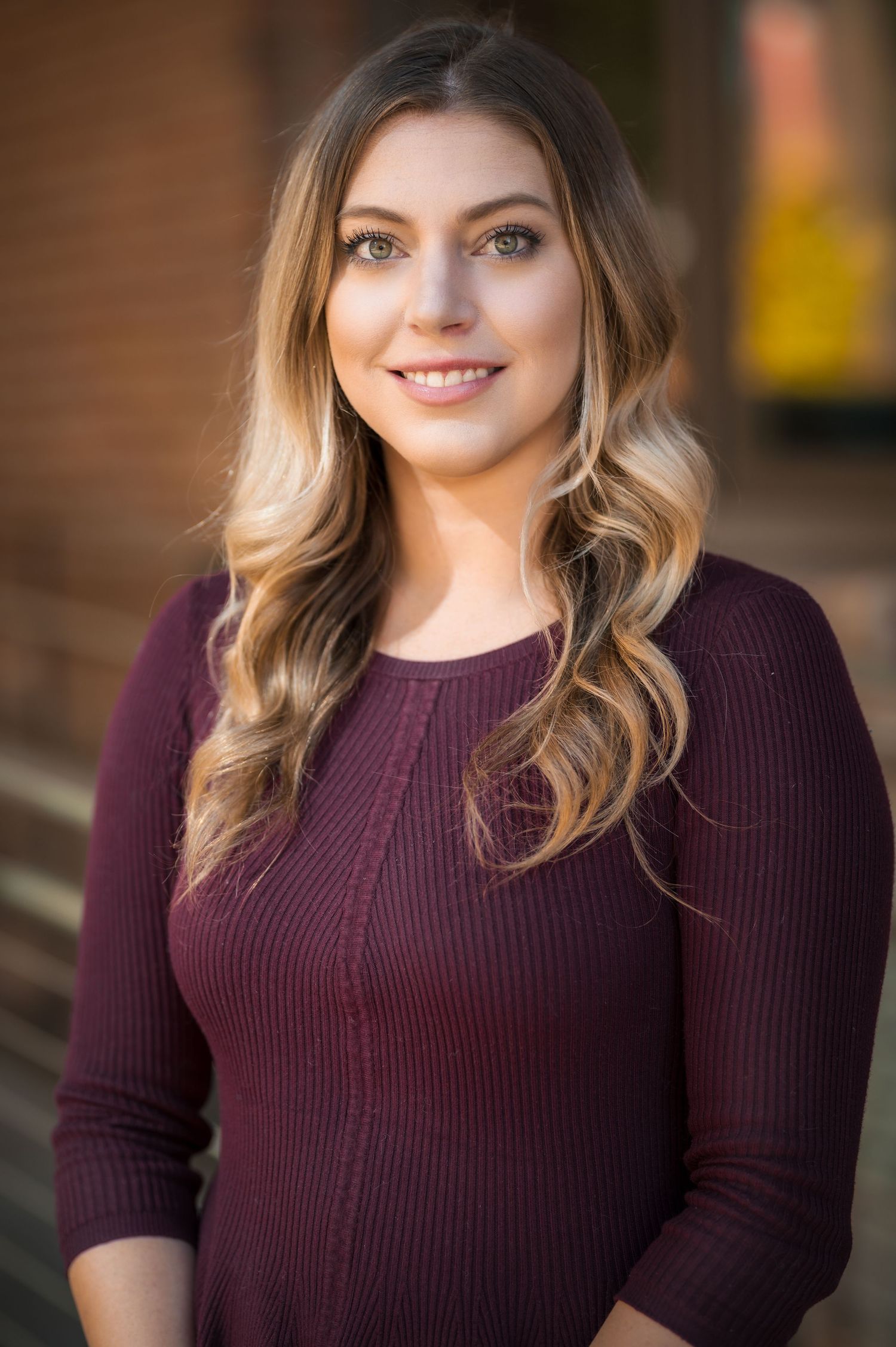Portrait series in burgundy sweater with natural makeup and highlighted hair against brick background.