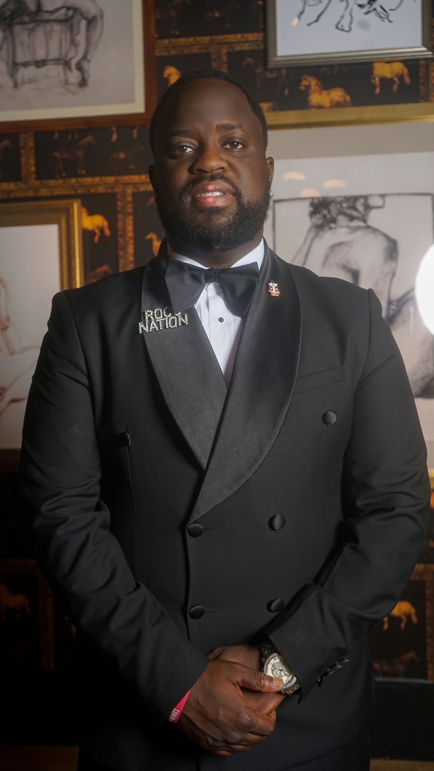 Man in black double-breasted tuxedo with bow tie and Roc Nation lapel pin poses confidently indoors.