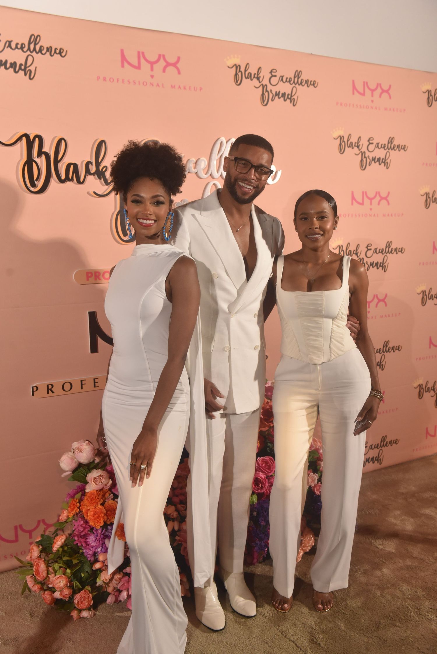 Three people in white outfits posing at NYX Professional Makeup Black Excellence Brunch event backdrop.