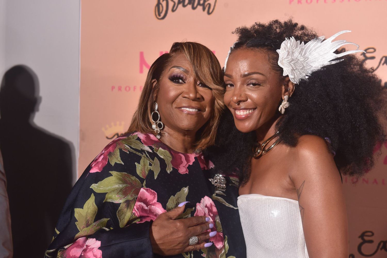 Two women smiling and posing together at a pink event backdrop, one in floral dress, other in white strapless top with hair accessory.