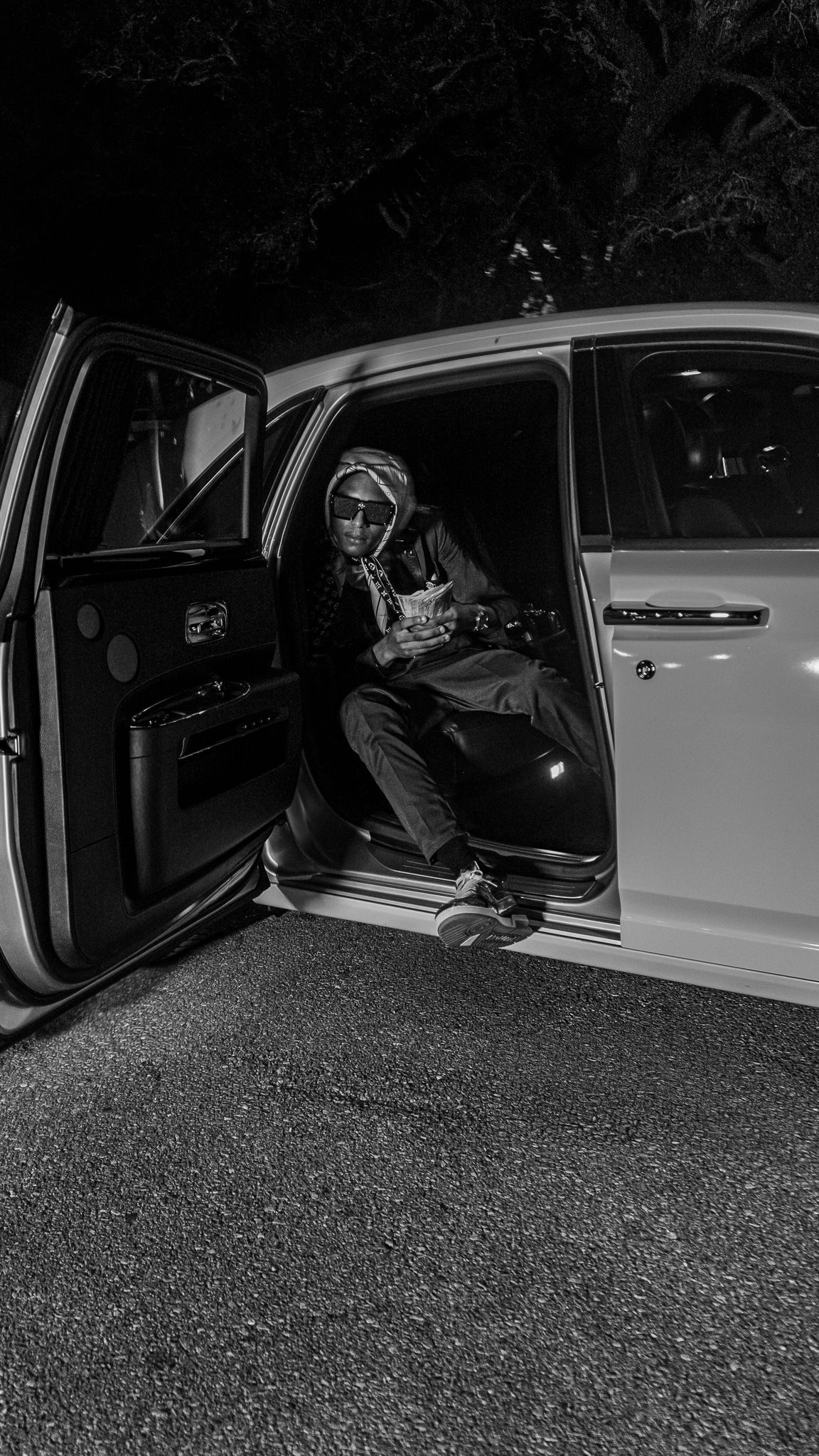 Black and white photo of a person sitting in an open car door at night, wearing sunglasses and casual clothing.