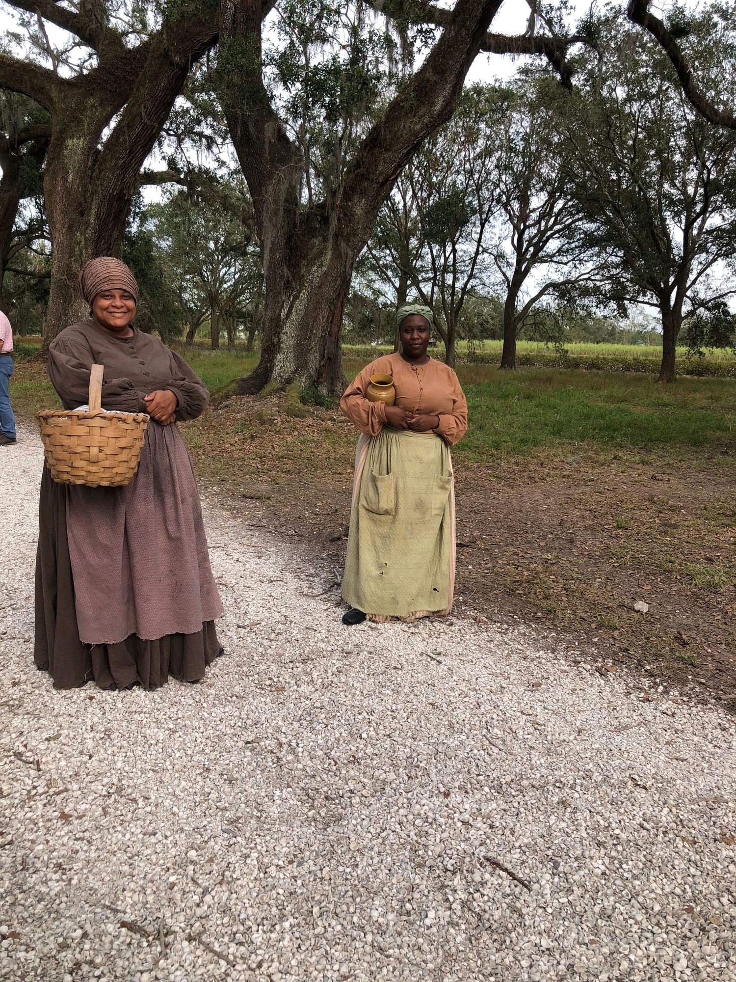 Two figures in long dresses stand on a gravel path surrounded by trees.