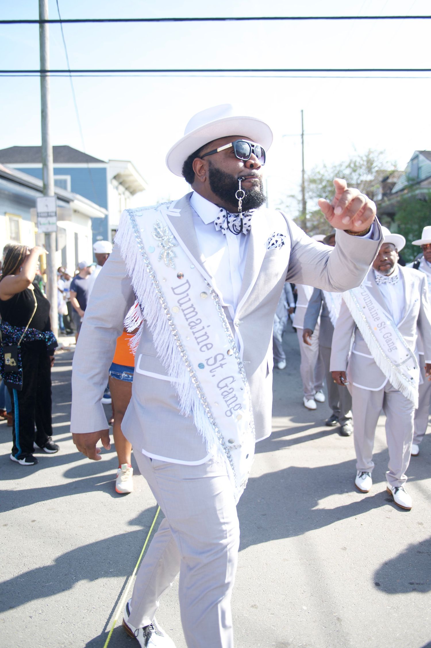 Parade participant in all-white suit with sash and hat leading a second line dance.