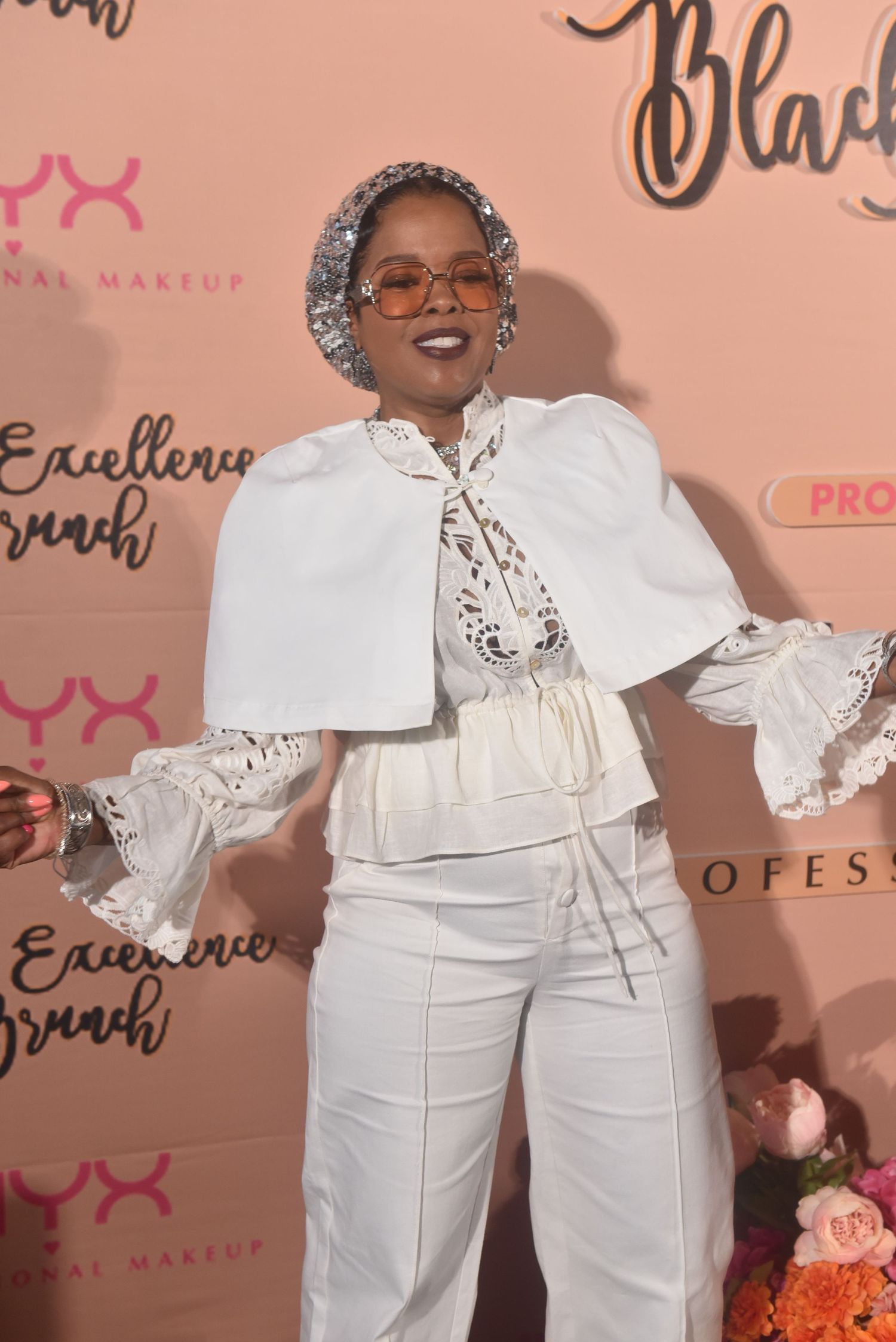 Woman in white cape outfit with sequin headscarf and orange glasses poses at NYX Professional Makeup Excellence Brunch.