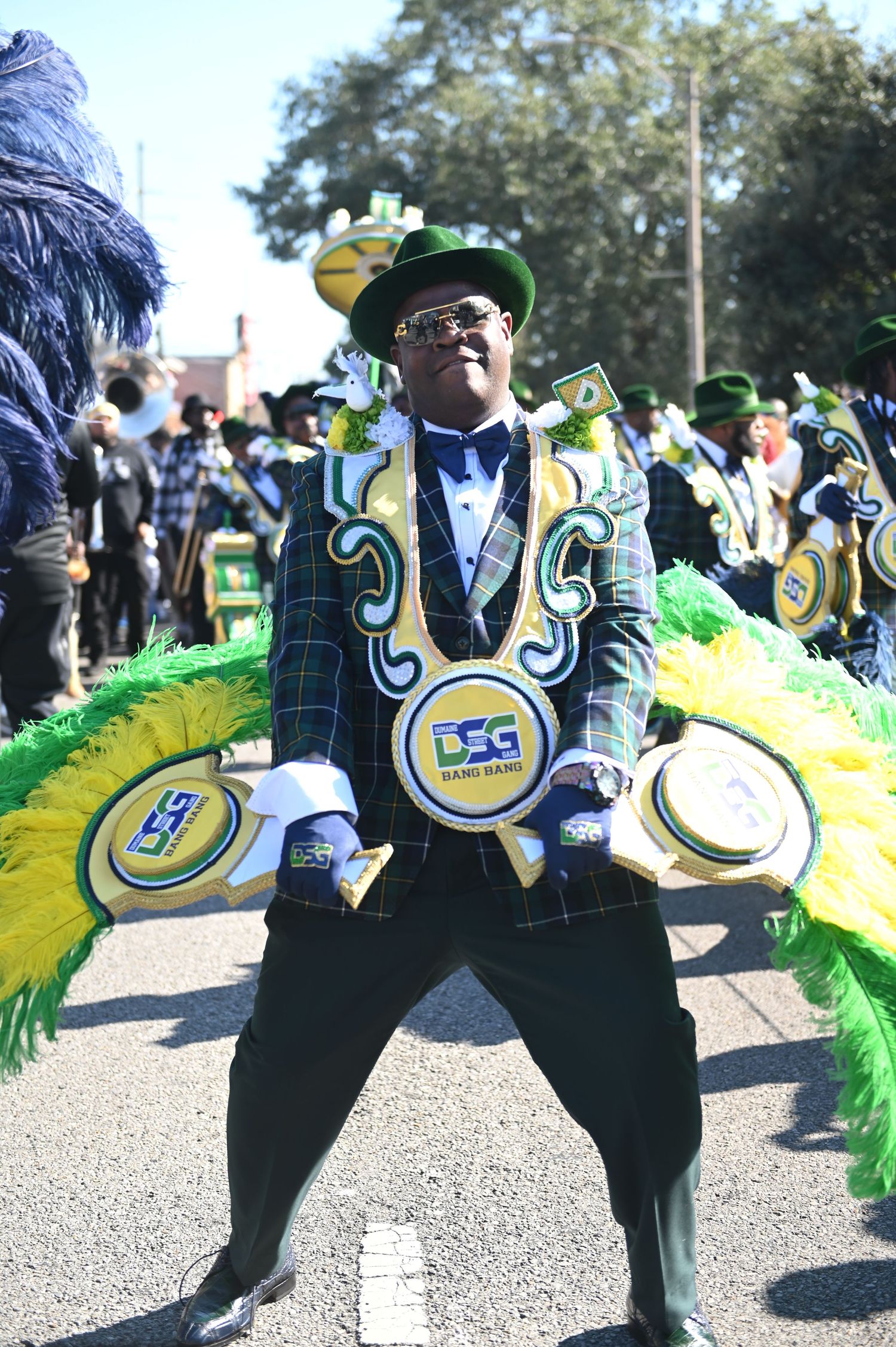 Performers in black and gold costumes dance in formation during a festive parade with bright green feathered decorations.