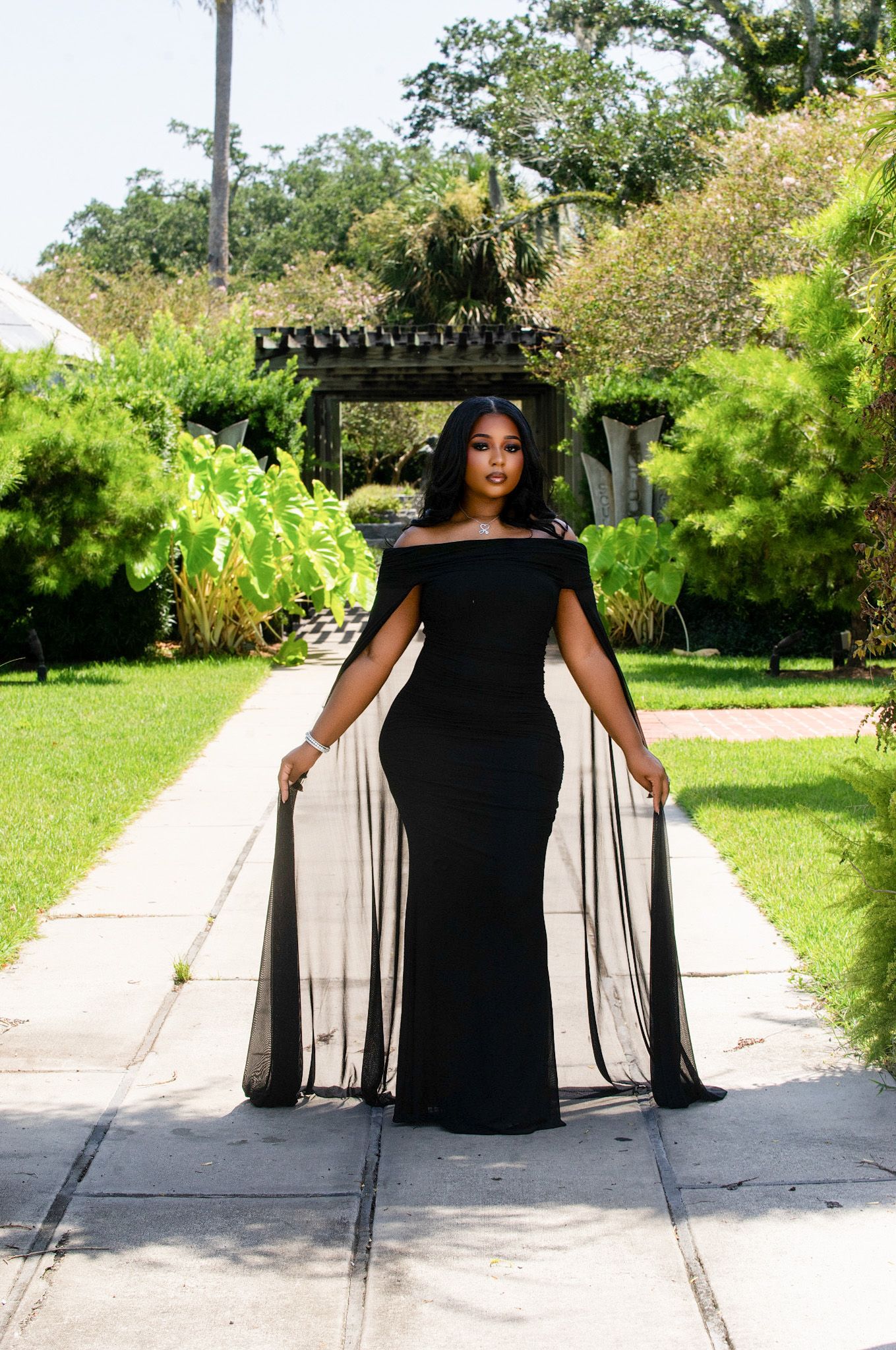 Woman in elegant black off-shoulder gown with sheer cape poses on garden pathway surrounded by lush greenery.