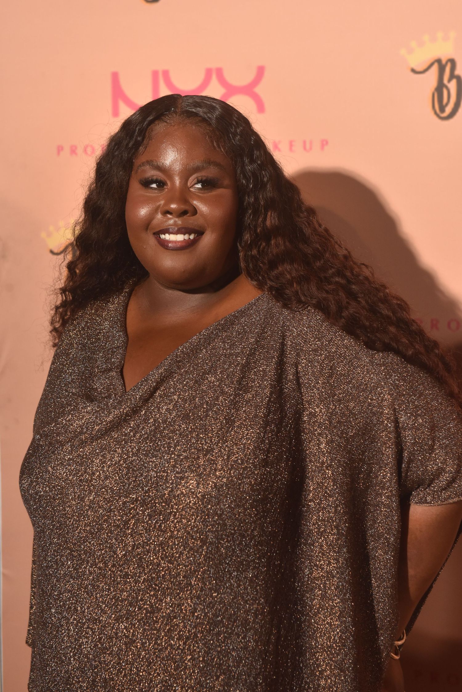 Woman in gold sparkly dress smiling at NYX Professional Makeup event on pink backdrop.