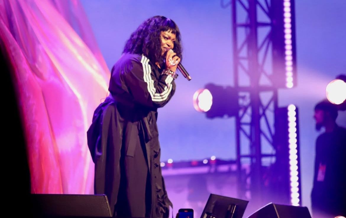 Performer in dark outfit singing on stage with purple lighting effects.