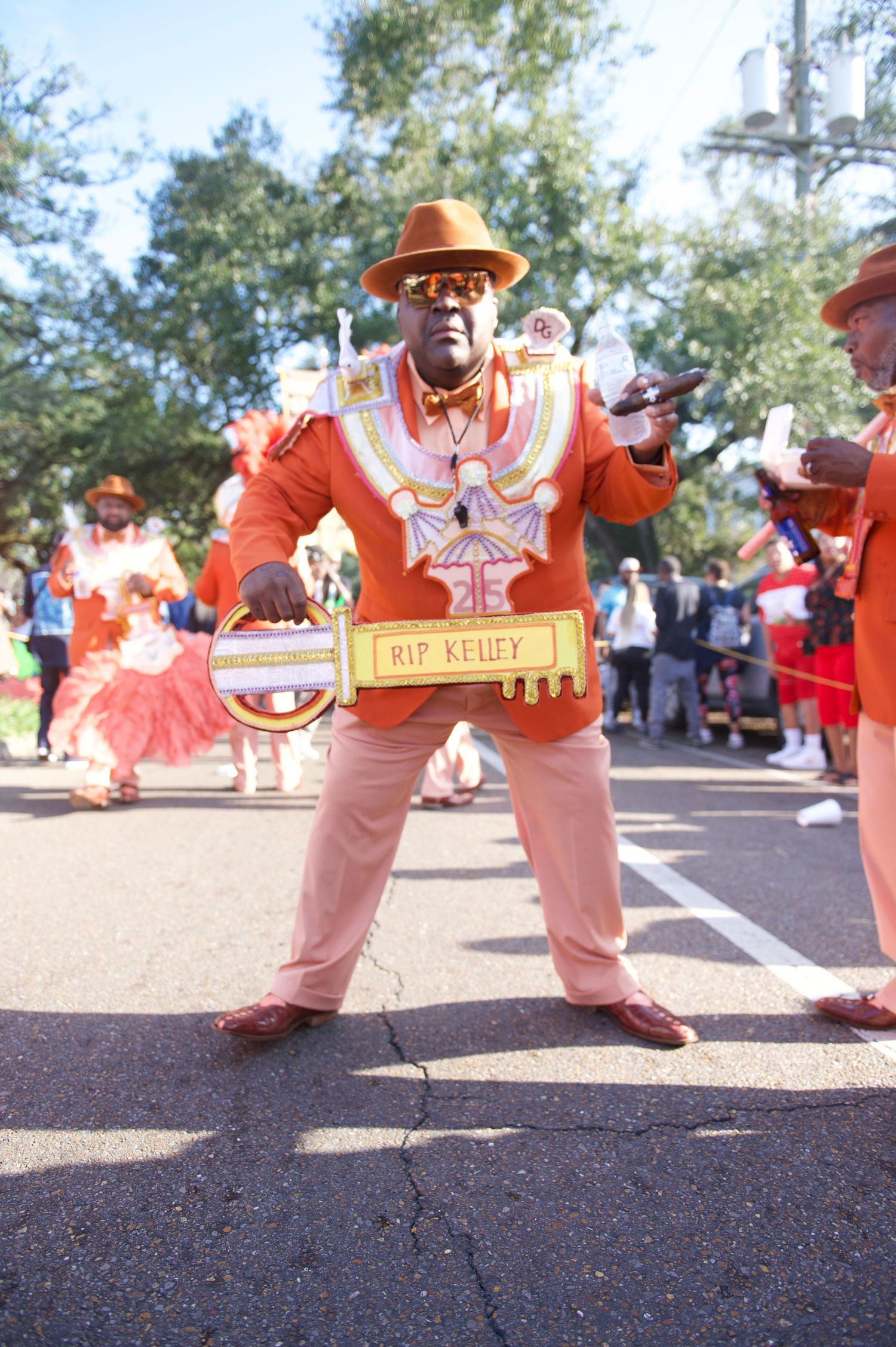 Performer in orange suit and hat carrying a decorative sign during a street parade.