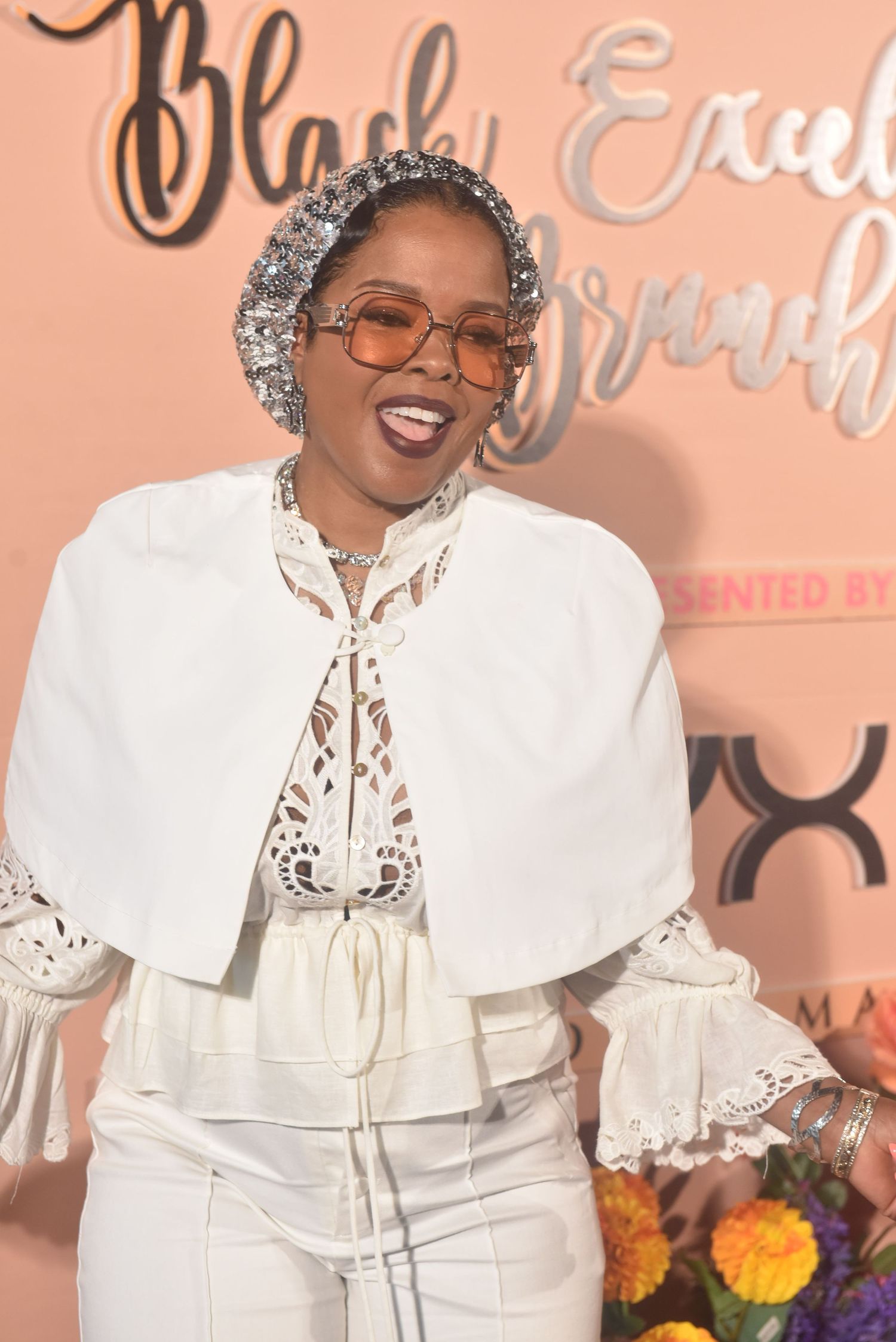 Woman in white outfit with silver glitter hat and orange sunglasses smiling at Black Excellence Brunch event.