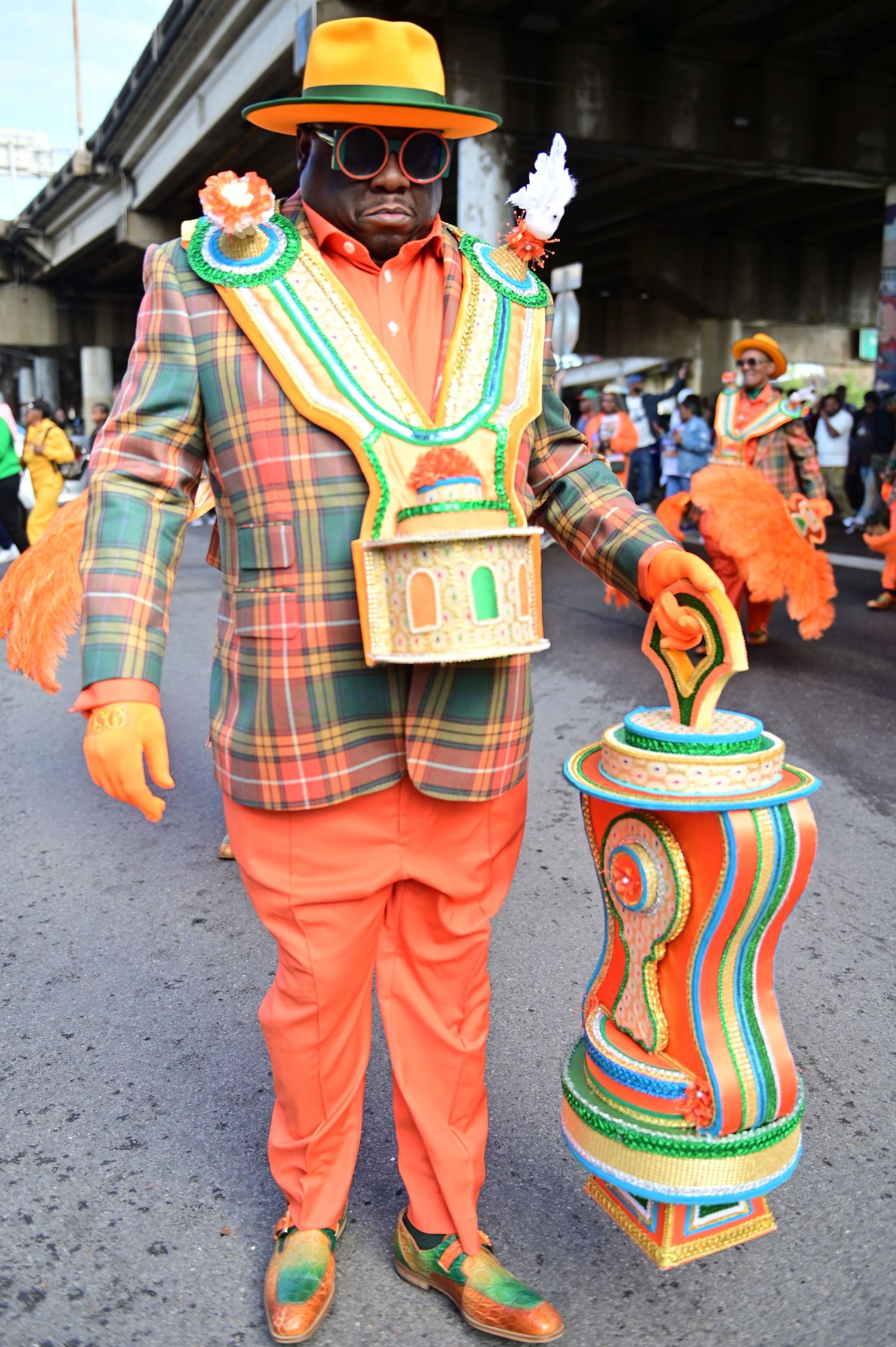 Costumed performer in orange and plaid outfit with decorative accessories poses on street.