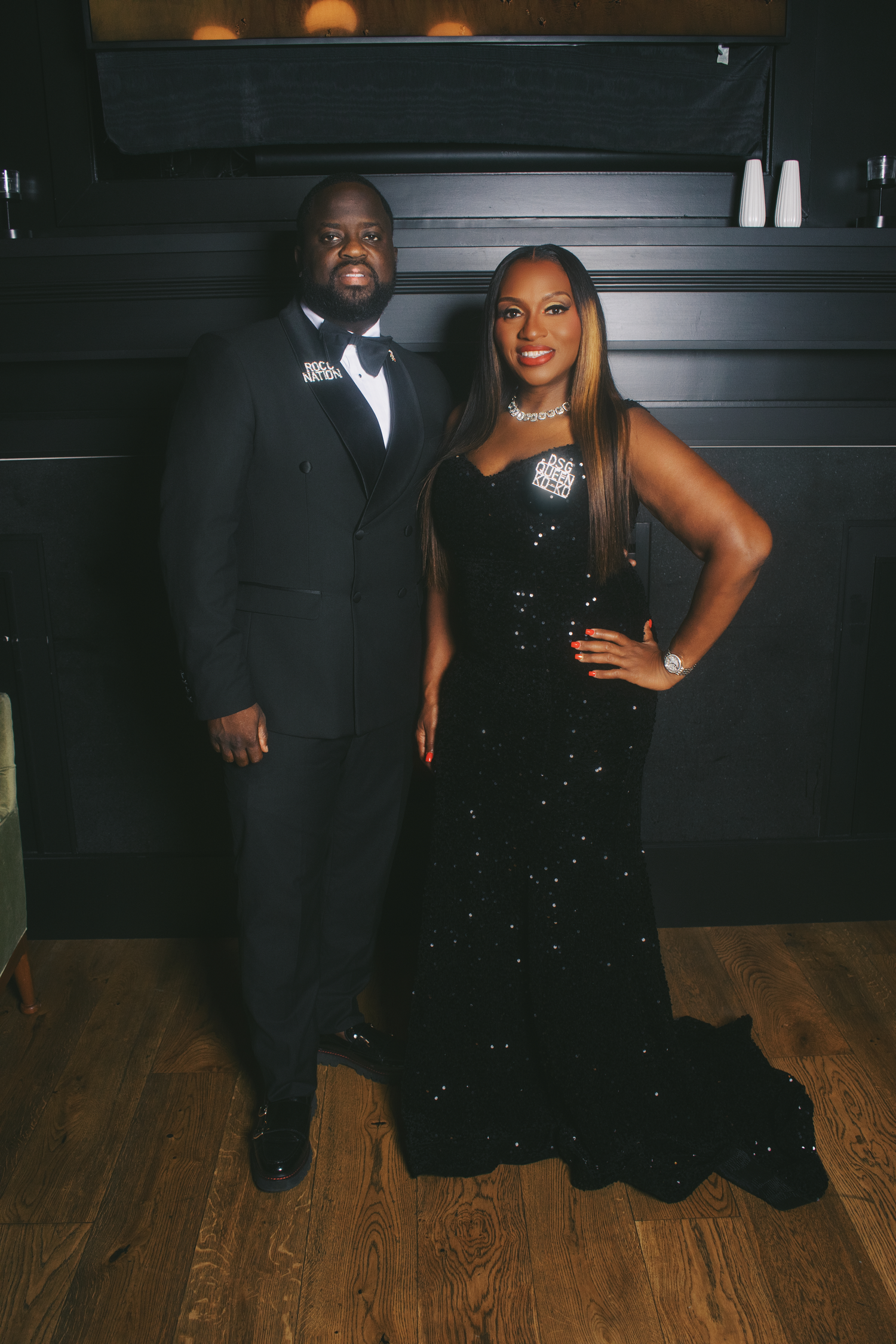 Elegant couple posing in matching black formal attire, man in tuxedo and woman in sequined gown, at a upscale event.