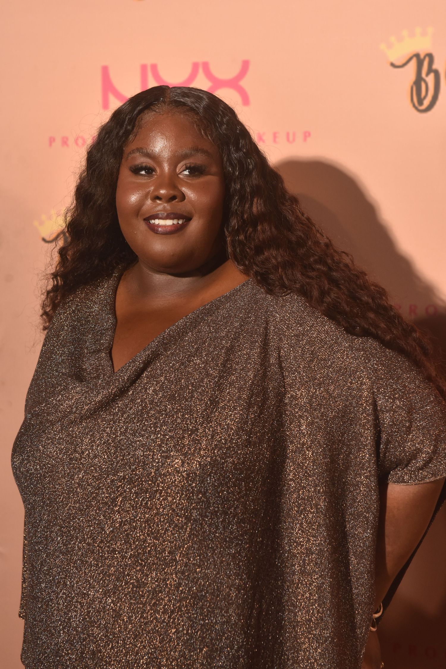 A smiling woman in a gold shimmer dress poses at a NYX Professional Makeup event backdrop.