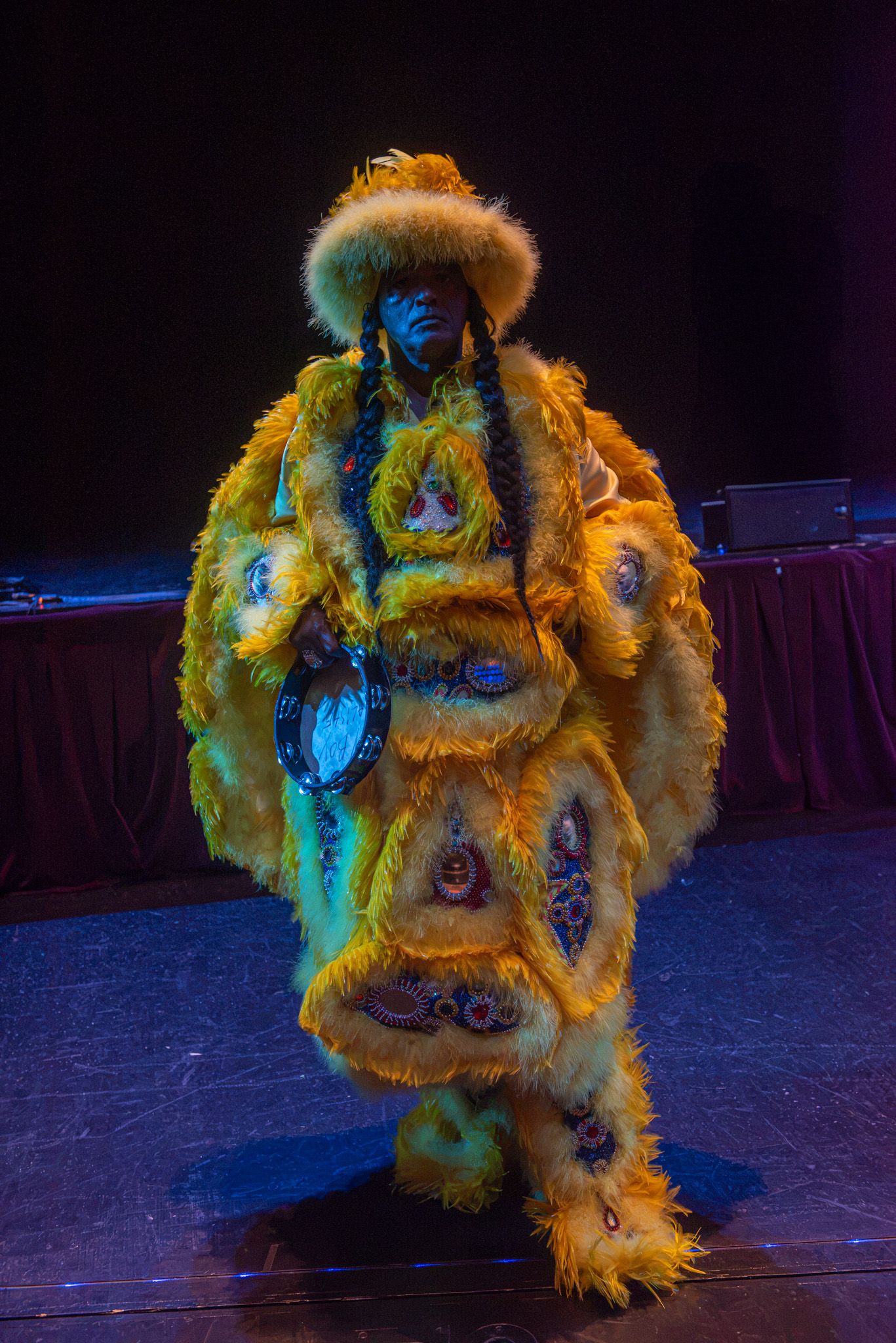 Performer in elaborate yellow fur costume with ornate decorations and matching hat stands on a dimly lit stage.